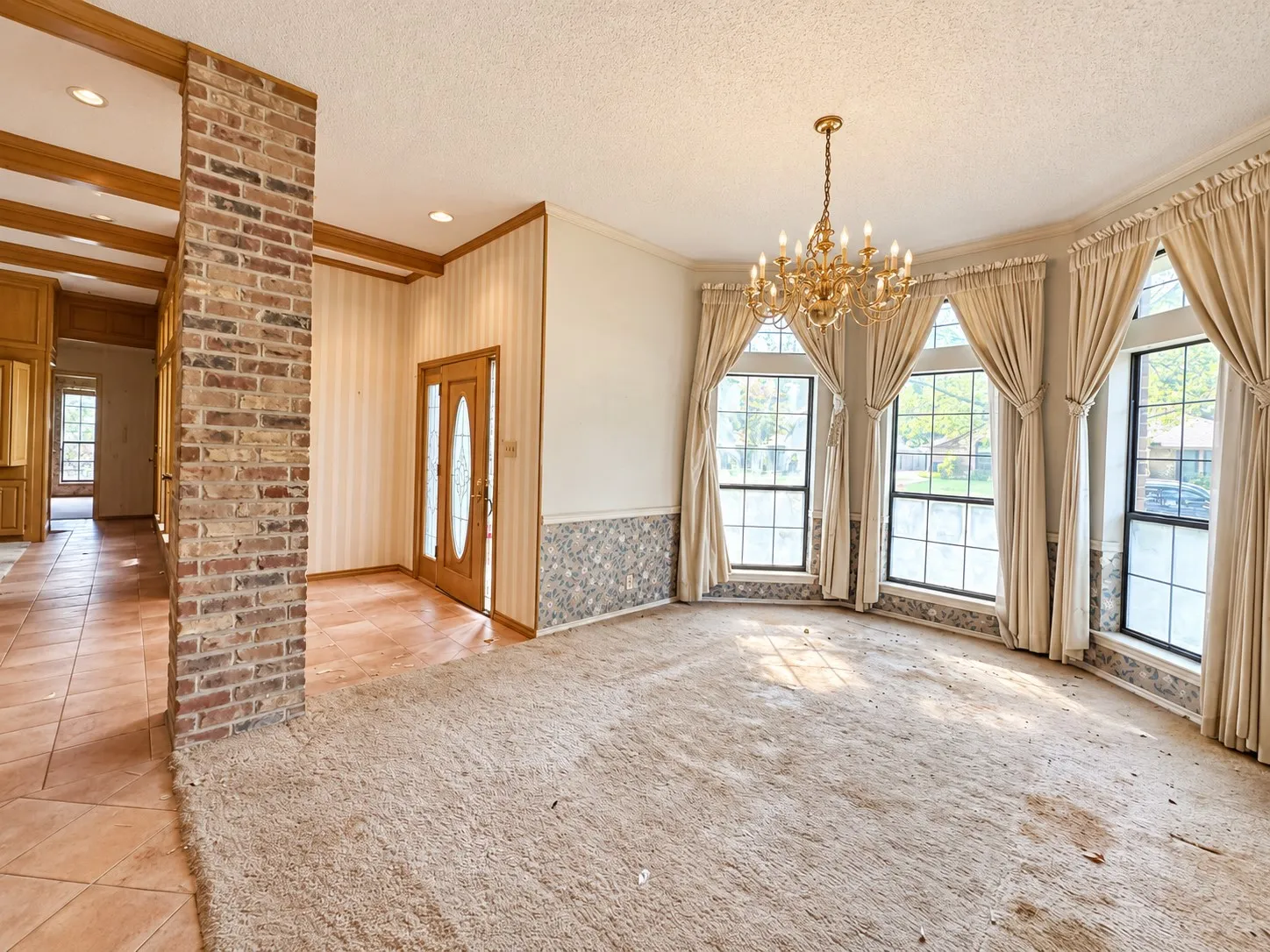 Bay window wall, exposed brick column, ornate chandelier, wood-paneled ceiling with recessed lighting, and a double entry door with glass inserts