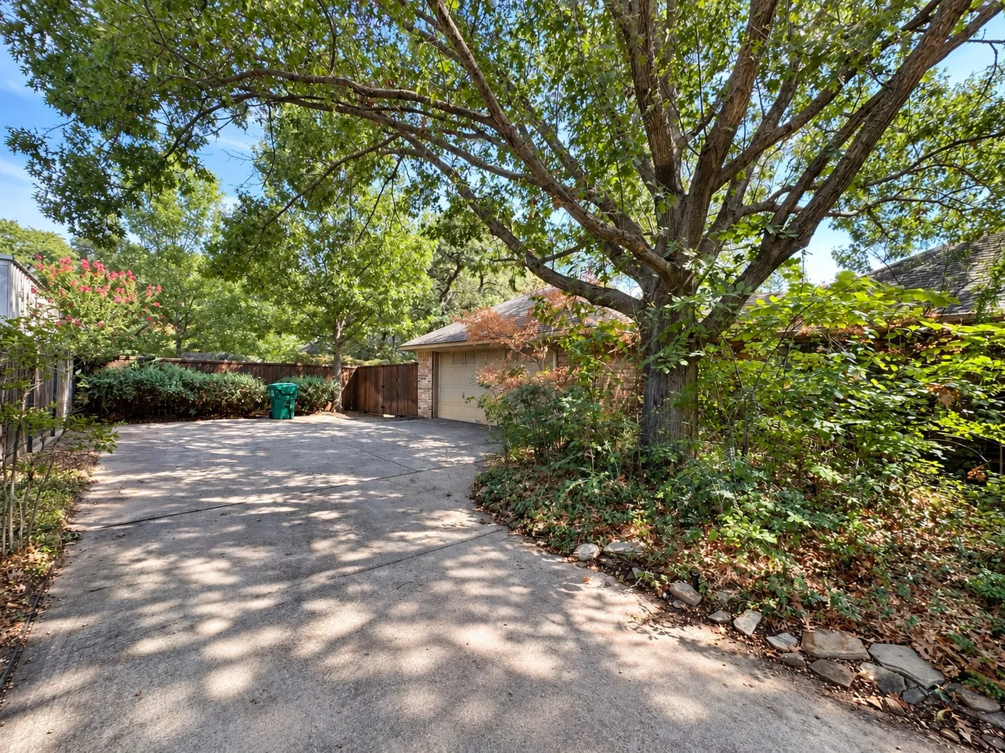 Concrete driveway framed by mature trees, a wooden fence, and lush landscaping, leading to a garage with a panel door