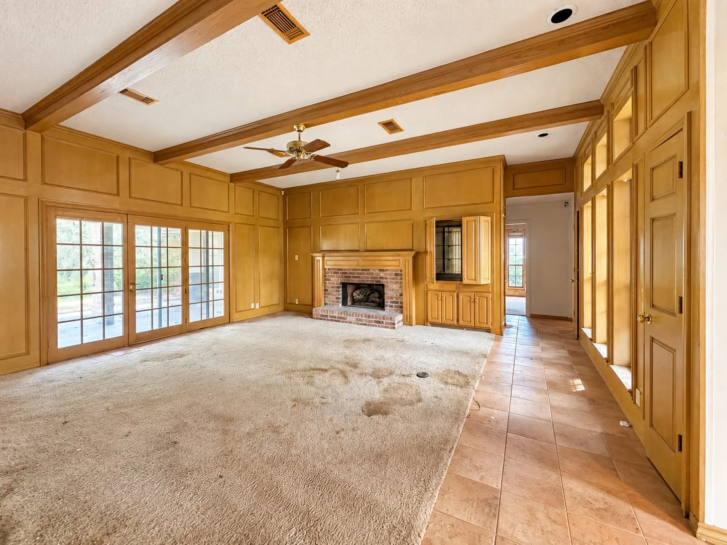 Wood-paneled walls with built-in cabinetry, exposed wood ceiling beams, brick fireplace with wood mantel, and multiple glass-paned doors