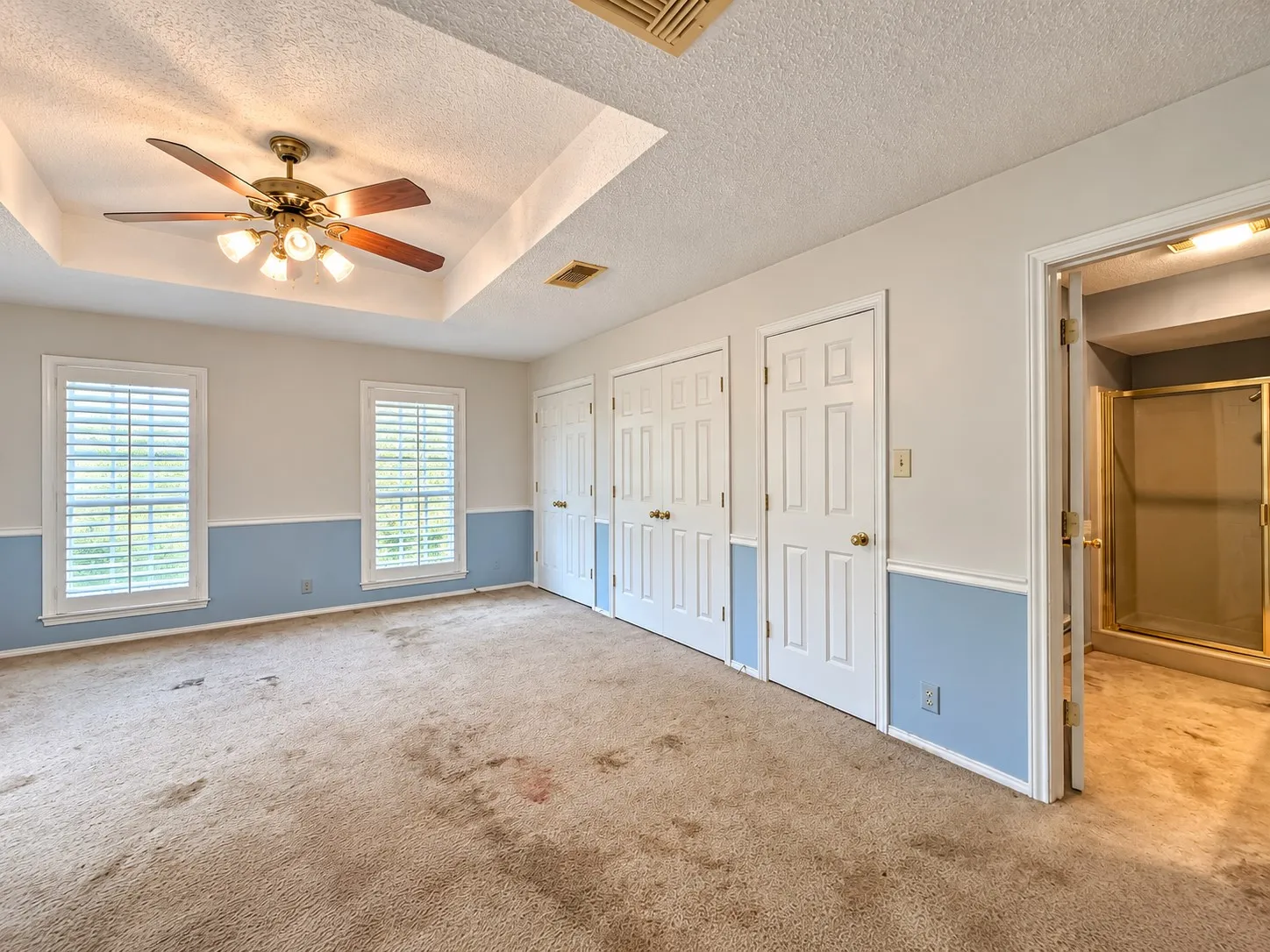 Tray ceiling with a ceiling fan, two windows with plantation shutters, multiple panel doors, and an open doorway to an adjoining room with a shower enclosure