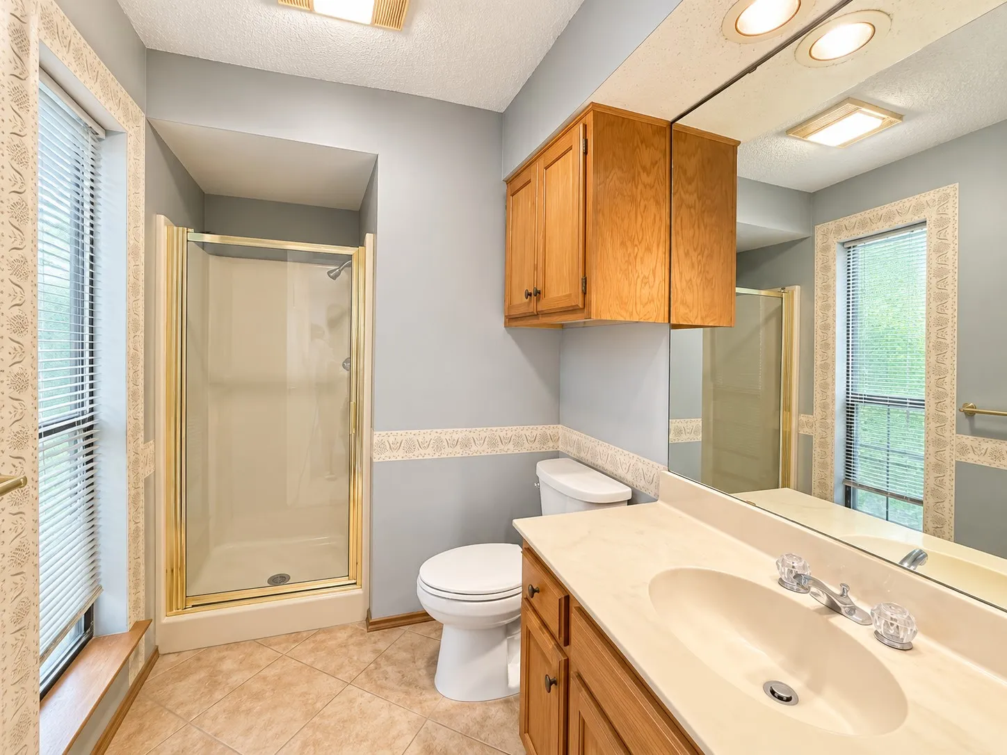 Bathroom featuring a shower enclosure with gold-tone framing, wood-finish vanity with an integrated sink, full-width mirrored wall, and overhead wood-finish cabinetry