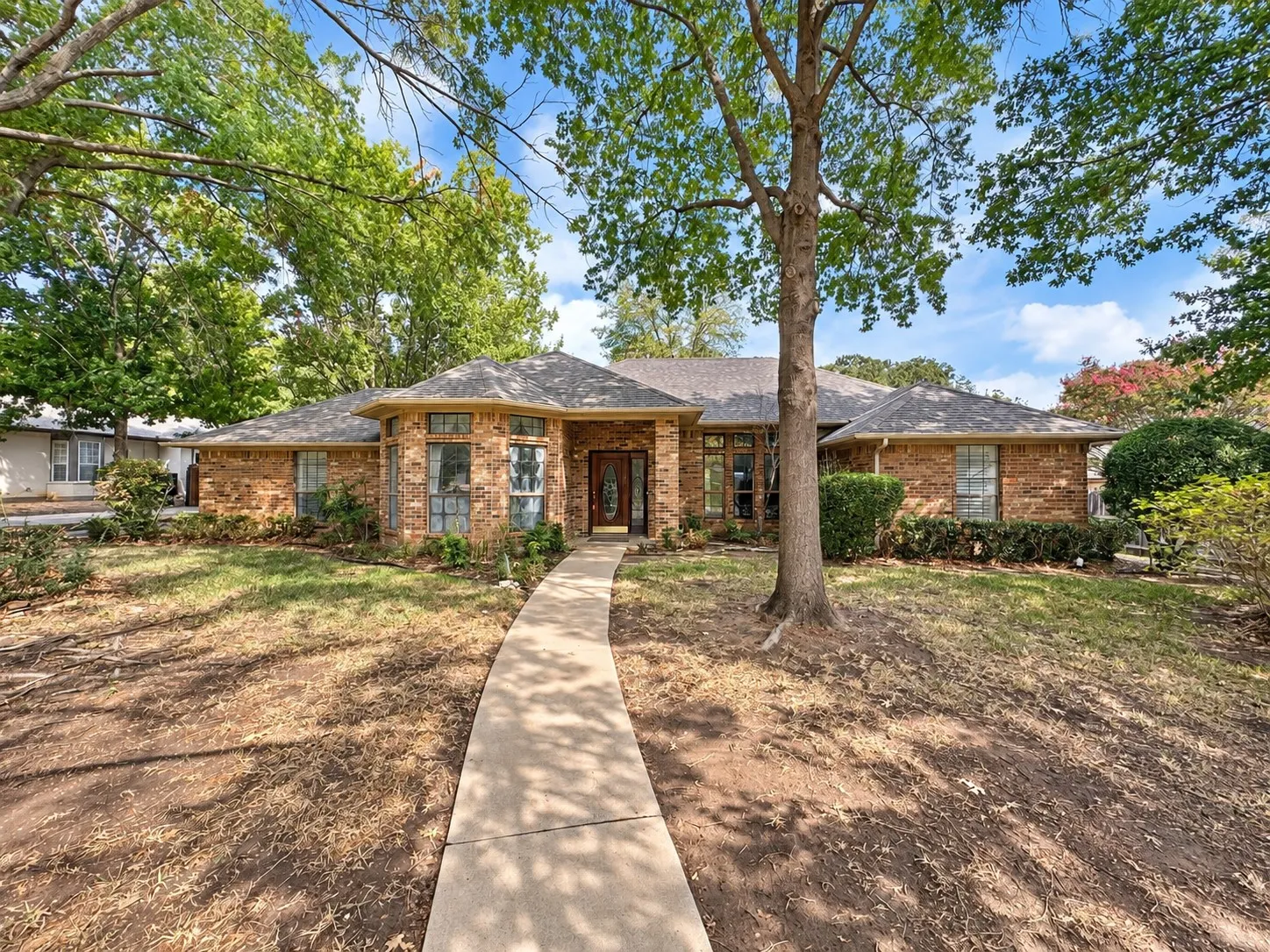 Brick exterior, mature trees, concrete pathway, bay windows, and a dark wood front door with decorative glass