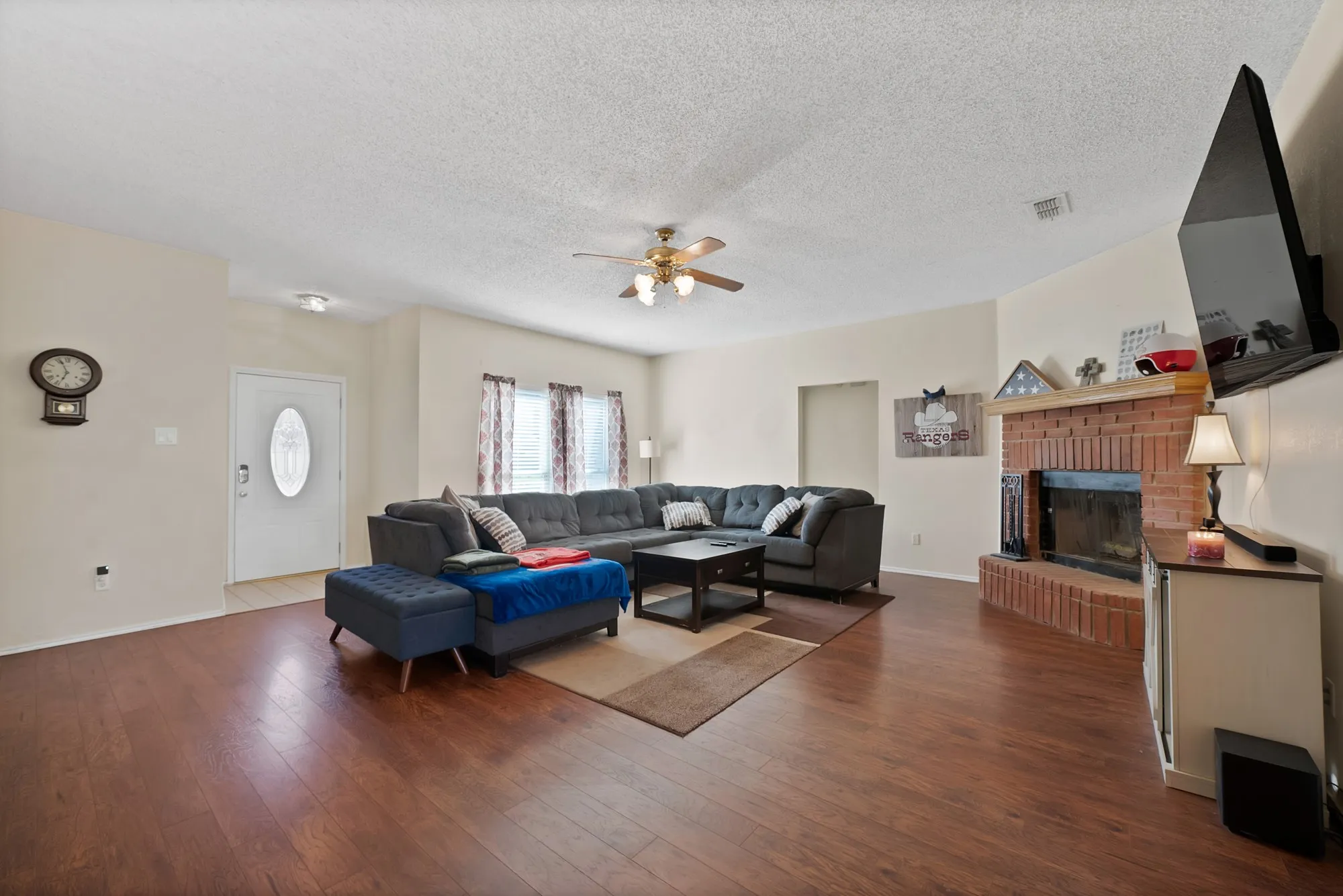 Wood-finish flooring throughout, featuring a brick fireplace with a mantel, a ceiling fan, and multiple windows