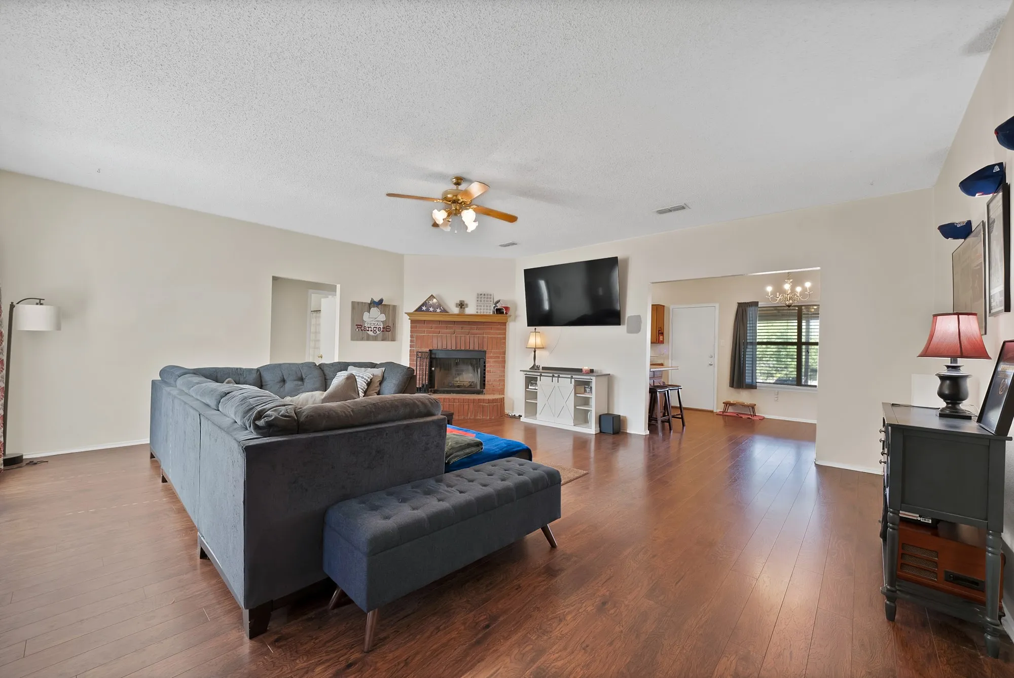 Expansive living space featuring wood-finish flooring, a corner brick fireplace, and a ceiling fan