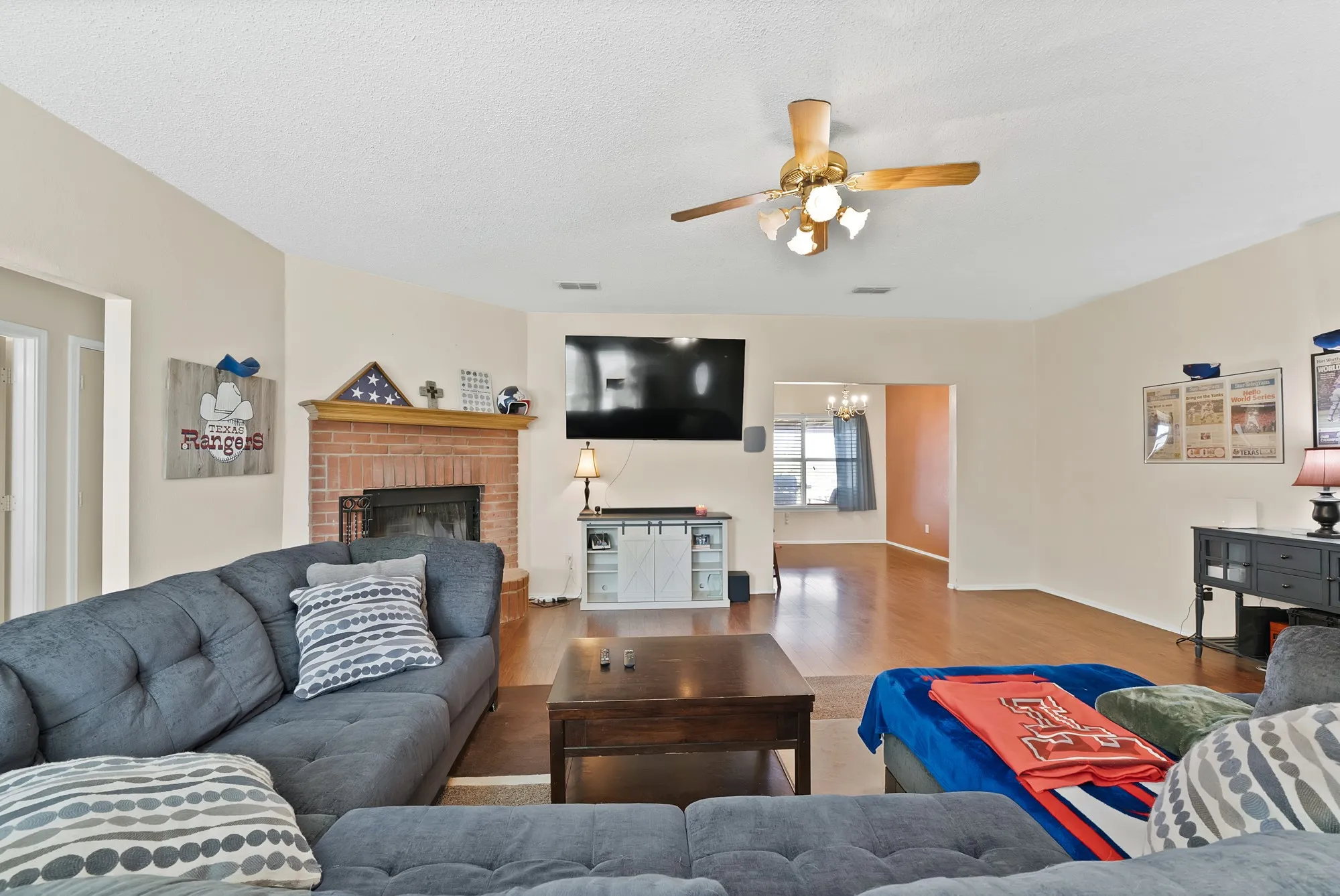 Warm living area featuring a brick fireplace with wood mantel, a ceiling fan with integrated lighting, wood-finish flooring, and an open doorway to another room