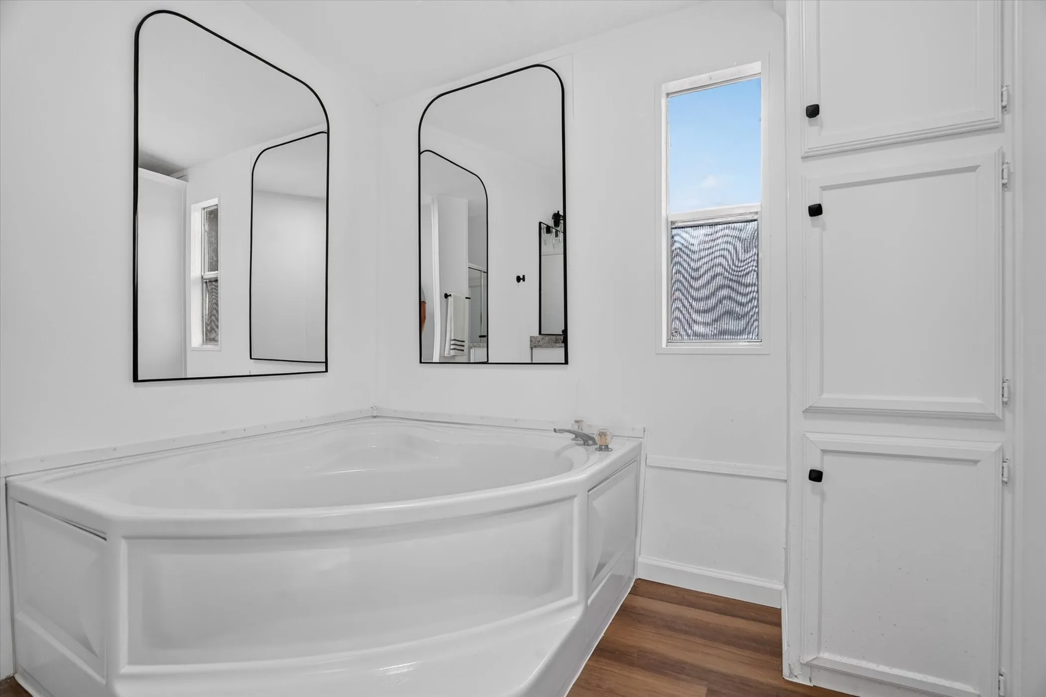 Corner soaking tub, dual arched mirrors, built-in storage cabinetry, wood-finish flooring, and a single window with privacy glass