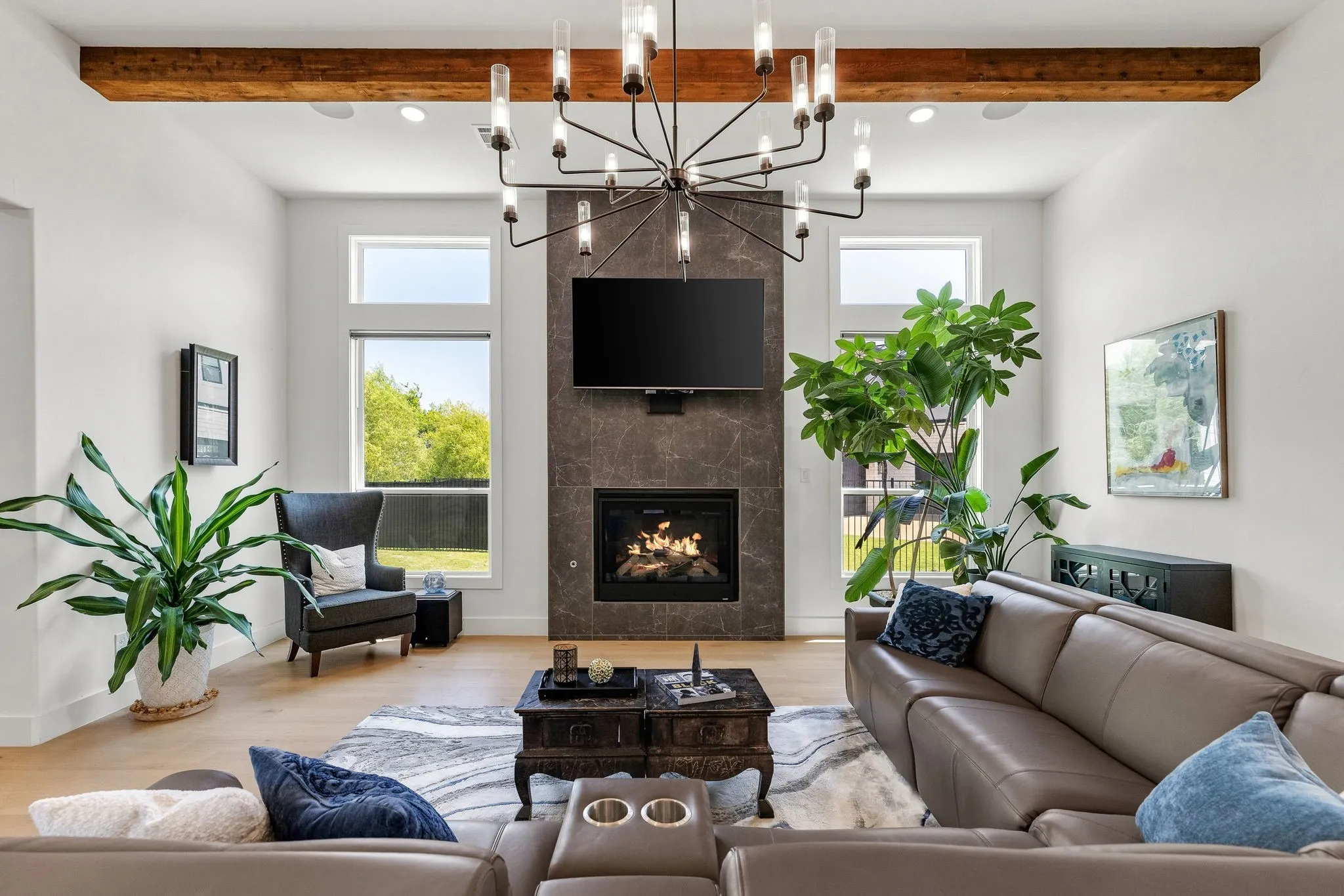 Light wood-finish flooring, a contemporary fireplace with a dark tile surround, a linear chandelier, and exposed wood beam ceiling accents
