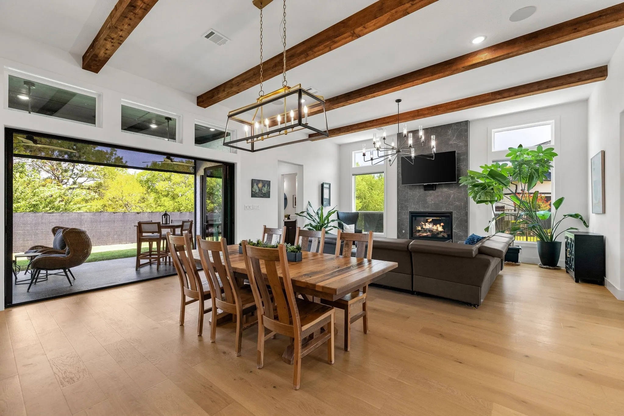 Vaulted ceiling with exposed wood beams, wide plank wood-finish flooring, full-height accordion glass doors, modern fireplace, and a stone-clad fireplace surround