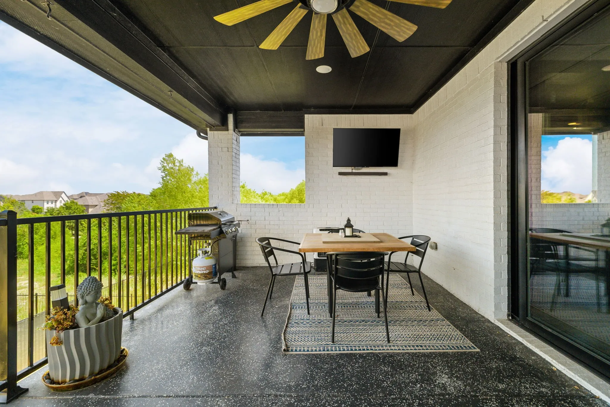 Covered balcony featuring a ceiling fan, white brick walls, black metal railing, and a dark, textured flooring surface