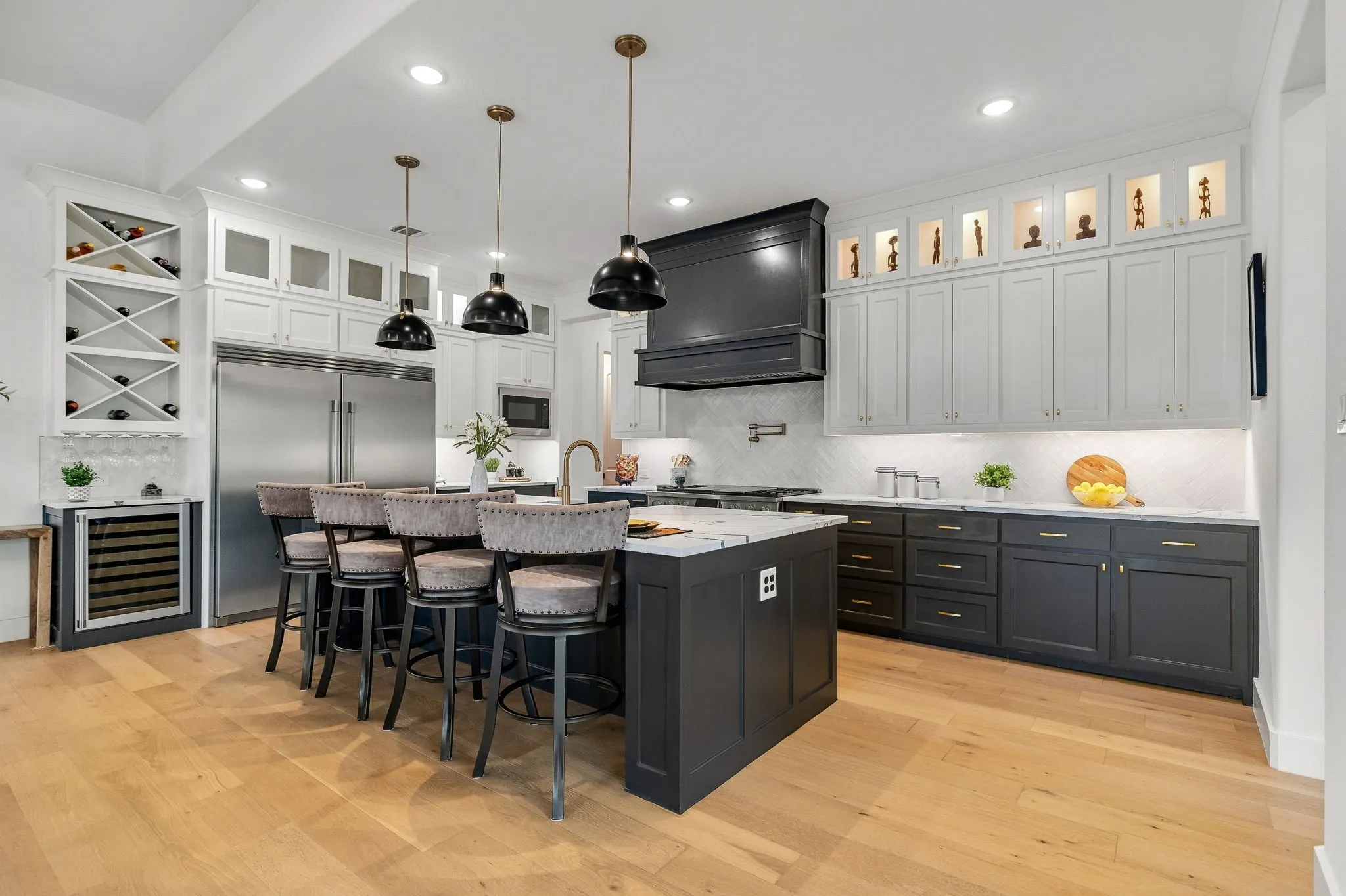 Gourmet kitchen featuring a large central island with seating, dark shaker-style cabinetry, white upper cabinetry with glass-front accents, light wood-finish flooring, and stainless steel appliances