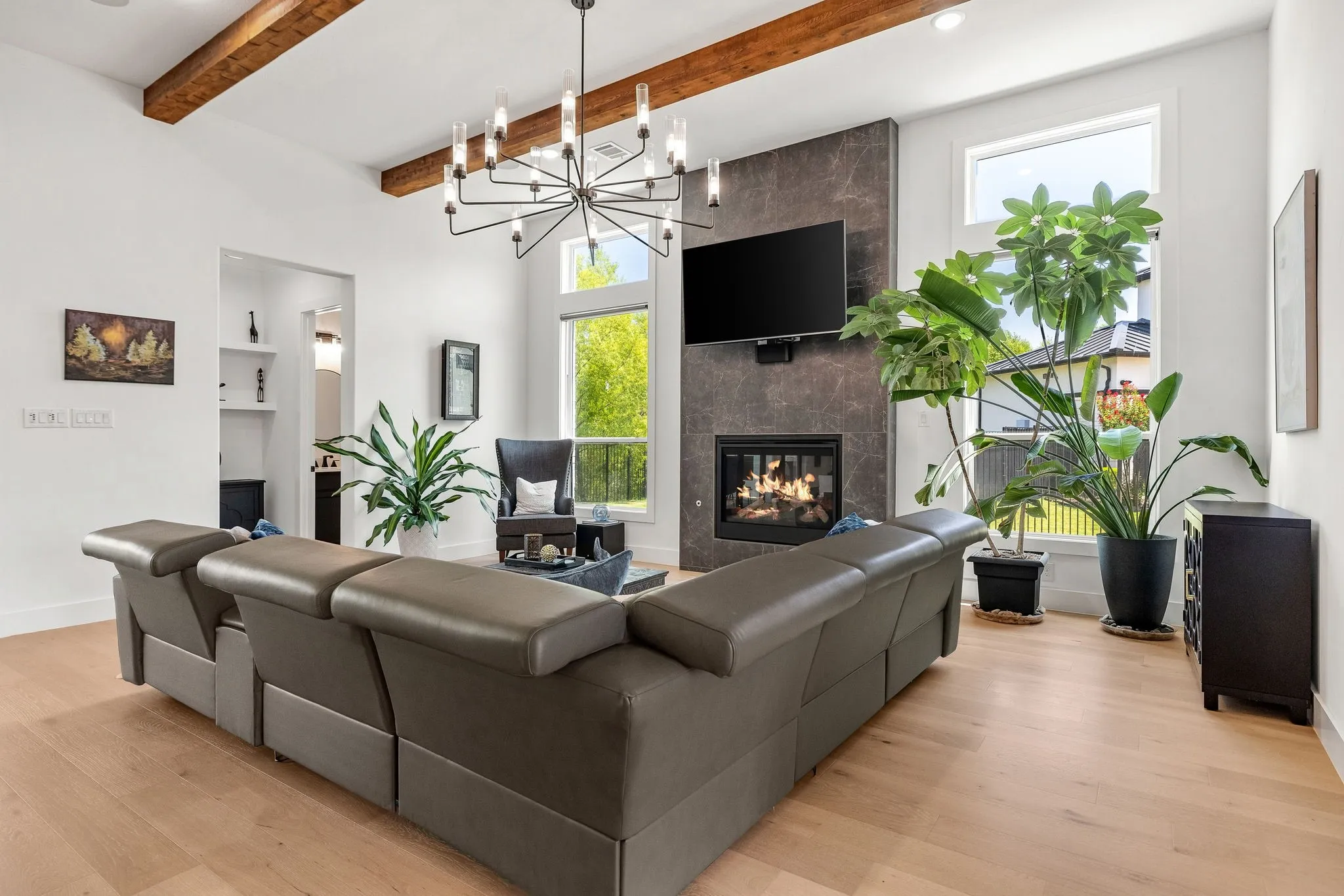 Spacious open-concept living area anchored by a gas fireplace with dark stone surround, complemented by exposed wood ceiling beams and a modern sputnik-style pendant fixture