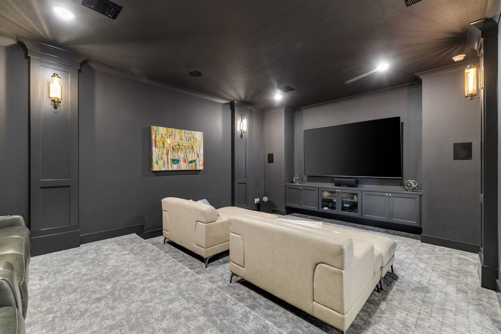 Dedicated media room featuring a large flat-screen television mounted above built-in cabinetry, recessed lighting throughout, dark gray walls, wall-mounted sconces, plush seating areas, and patterned area flooring