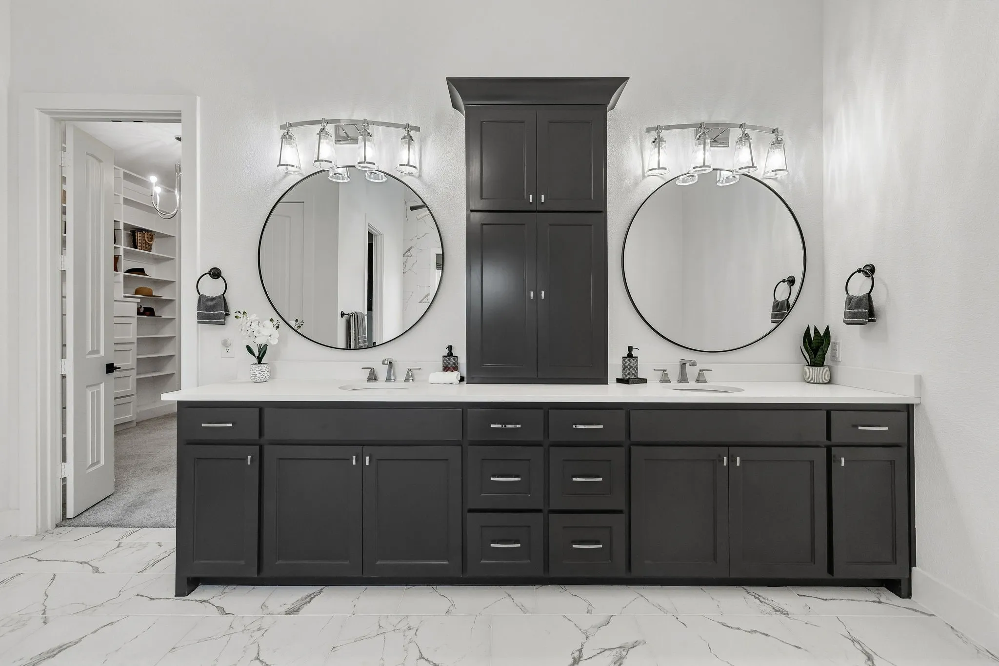 Luxurious dual-vanity bathroom featuring two circular framed mirrors with integrated lighting, extensive black cabinetry with multiple drawers and storage, polished stone flooring with linear patterning, chrome faucetry, and a tall black linen cabinet