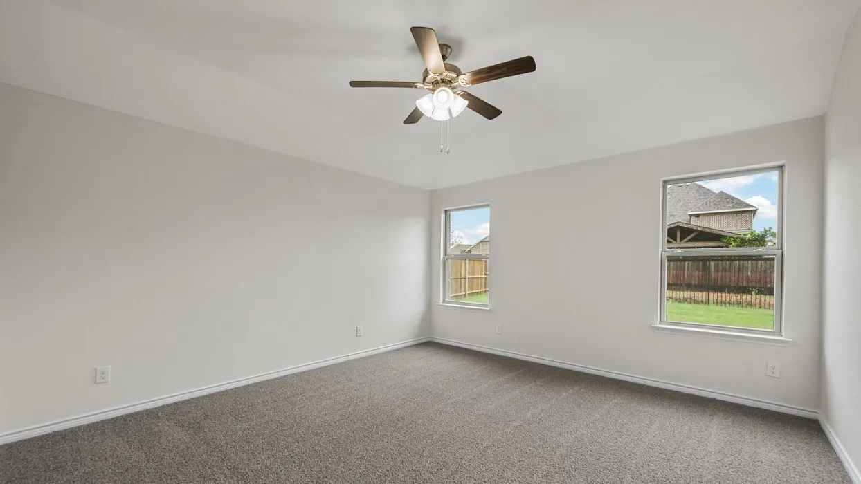 Bright and spacious bedroom showcasing neutral carpet flooring, dual windows with natural light, ceiling fan with integrated lighting, and fresh white walls