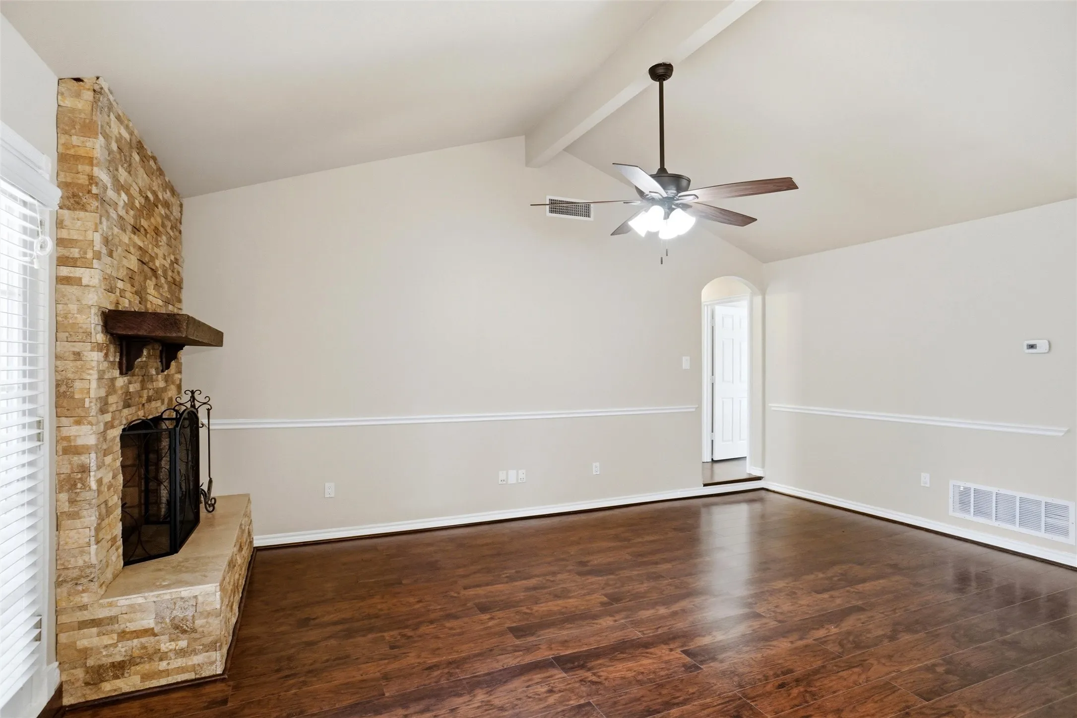 Spacious room with rich wood-finish flooring, exposed brick fireplace with hearth and mantel, ceiling fan with integrated lighting, and neutral two-tone wall treatment