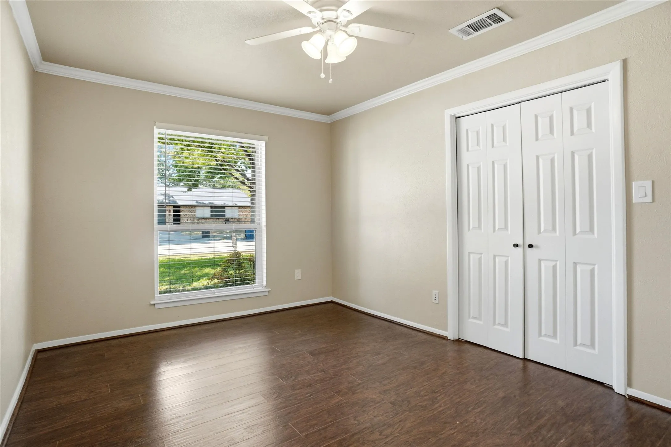 Wood-finish flooring, white bi-fold closet doors, crown molding, white ceiling fan with light, and a window with blinds