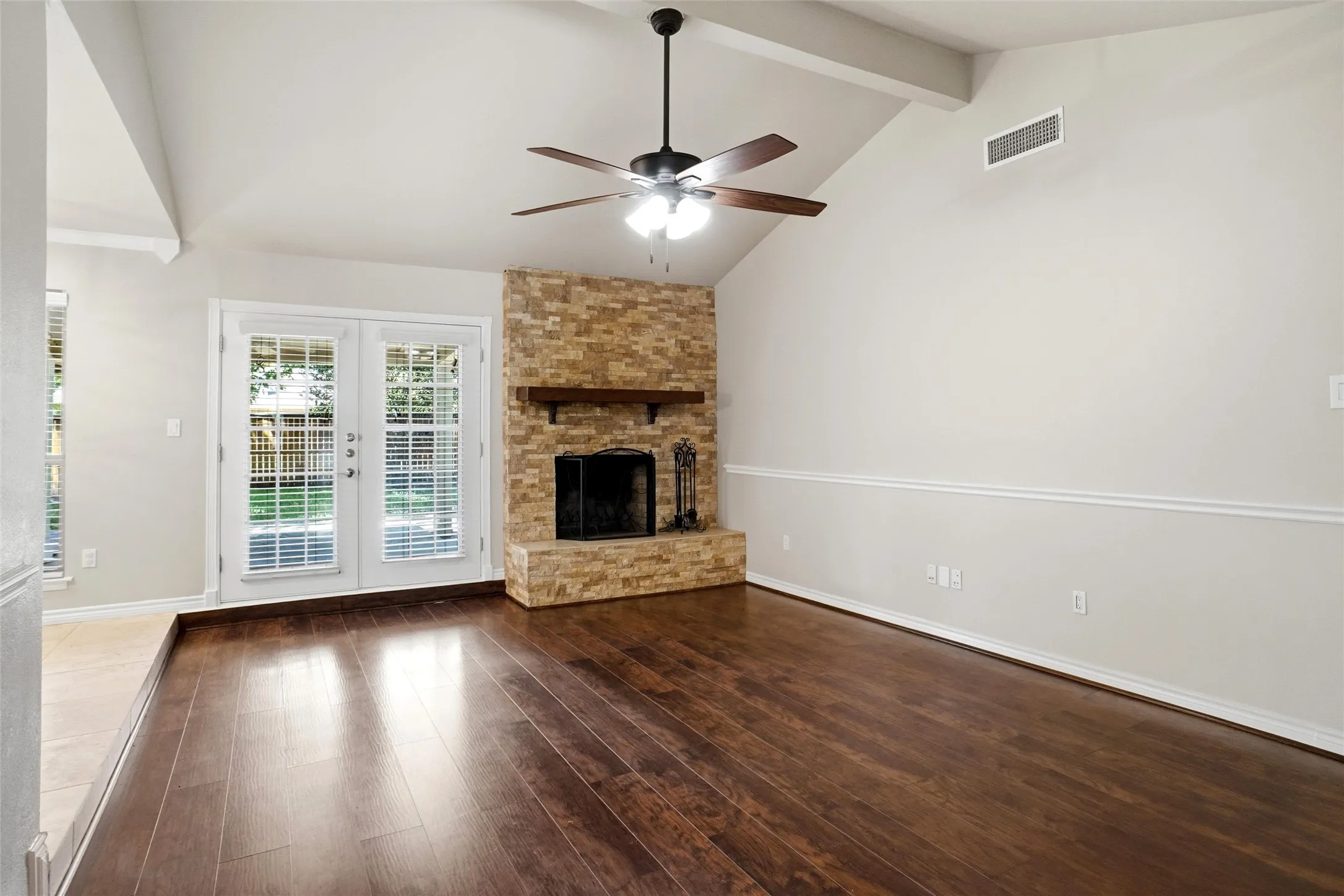 Vaulted ceiling with a ceiling fan, stone-clad fireplace with a wood mantle, rich wood-finish flooring, and double French doors opening to the exterior