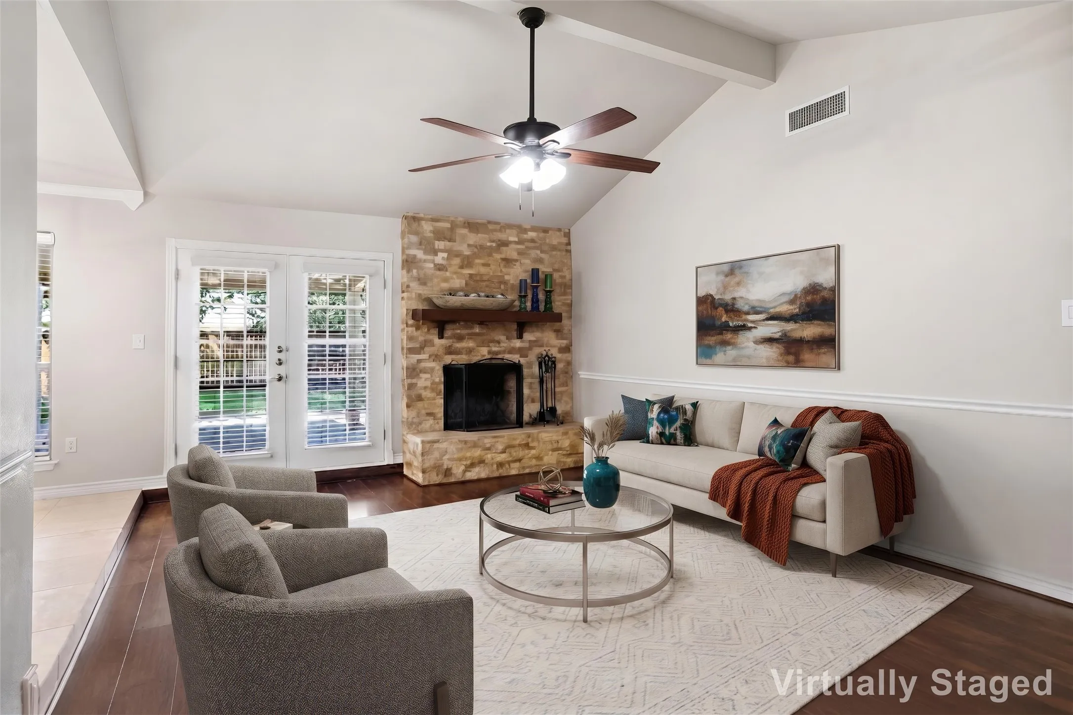 Vaulted ceiling with exposed beams, wood-finish flooring, stone-clad fireplace with wood mantel, French doors with integrated blinds, and a ceiling fan with integrated lighting