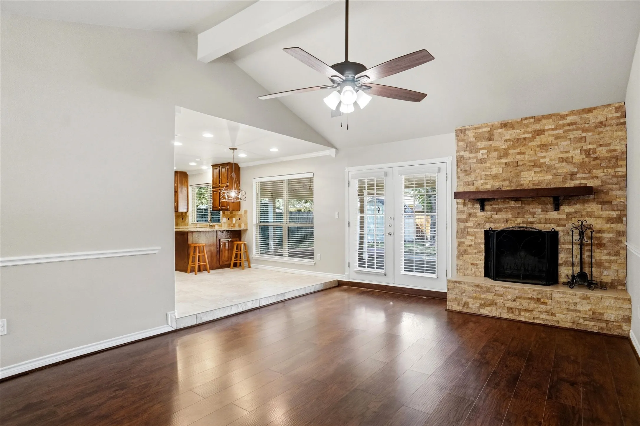 Vaulted ceiling with a ceiling fan, wood-finish flooring, stone-clad fireplace with a dark wood mantel, and double doors providing outdoor access