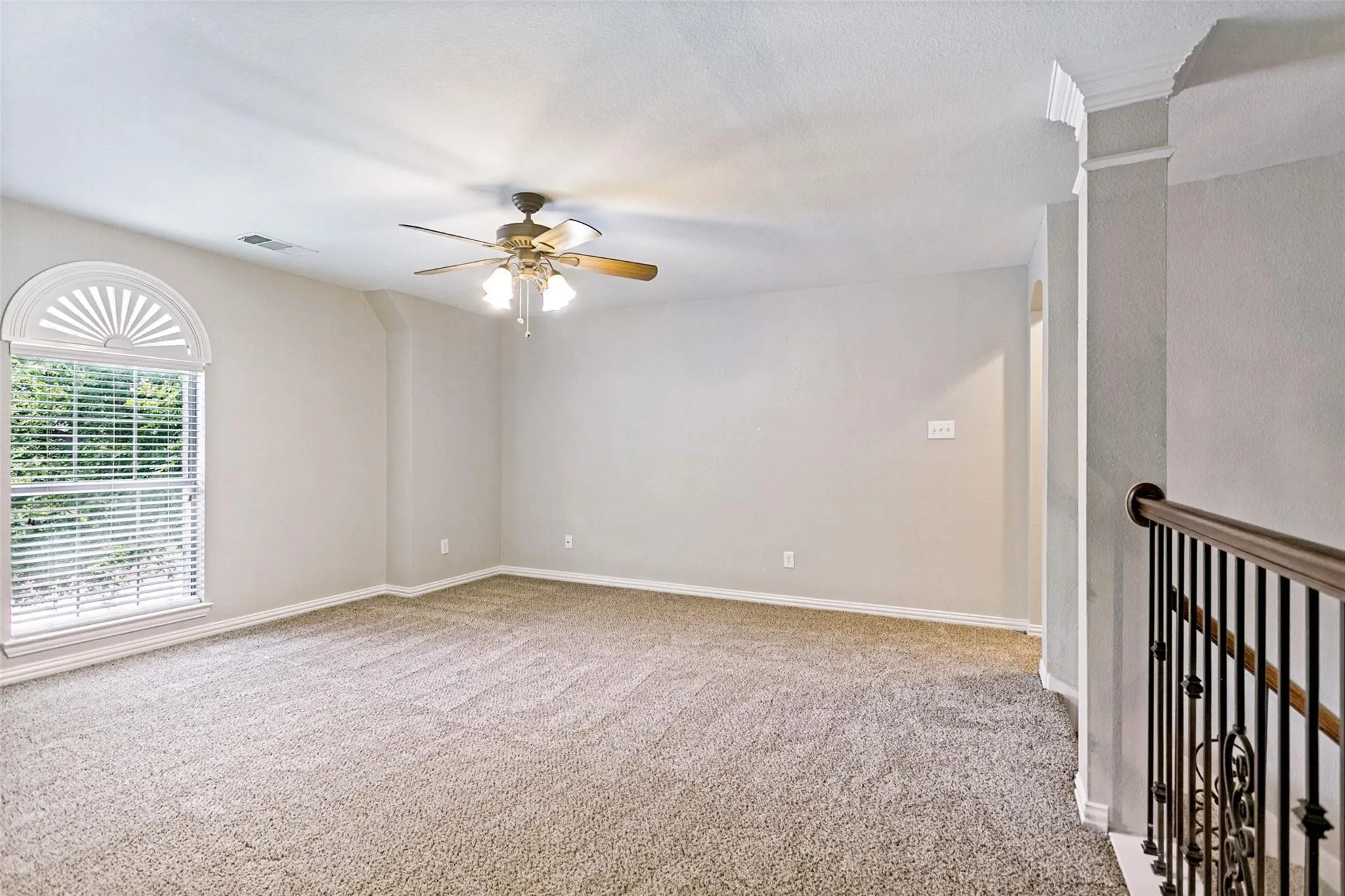 Arched window with louvered blinds, textured carpeting, ceiling fan with light fixture, decorative metal balusters, and crown molding