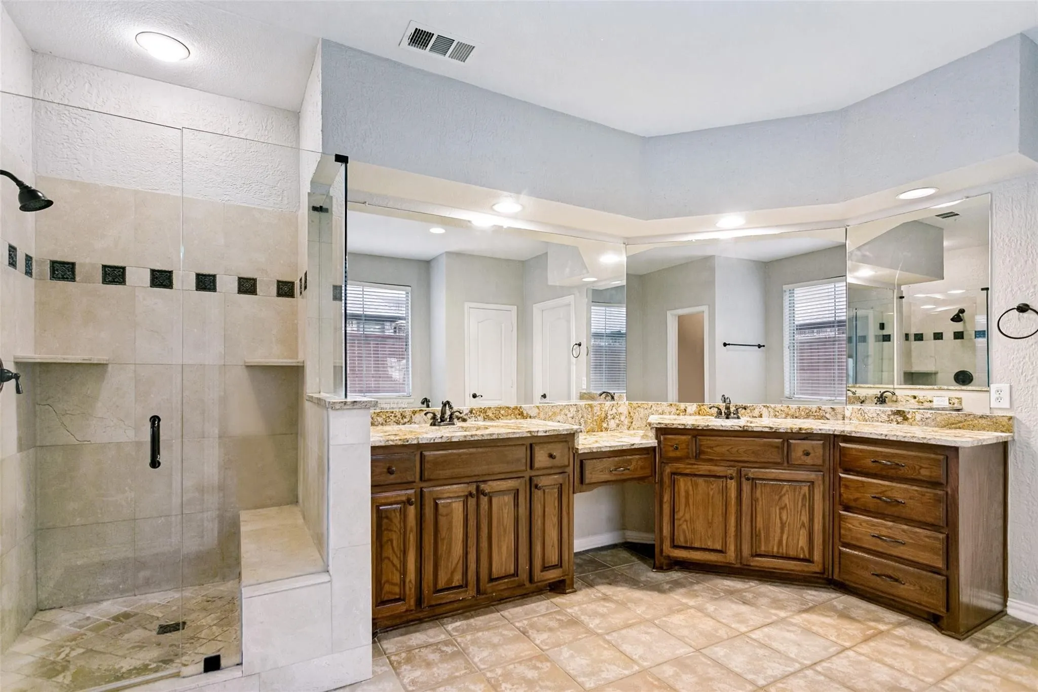 Spacious bathroom featuring a glass-enclosed shower with built-in bench, dual vanity with undermount sinks, and granite countertops
