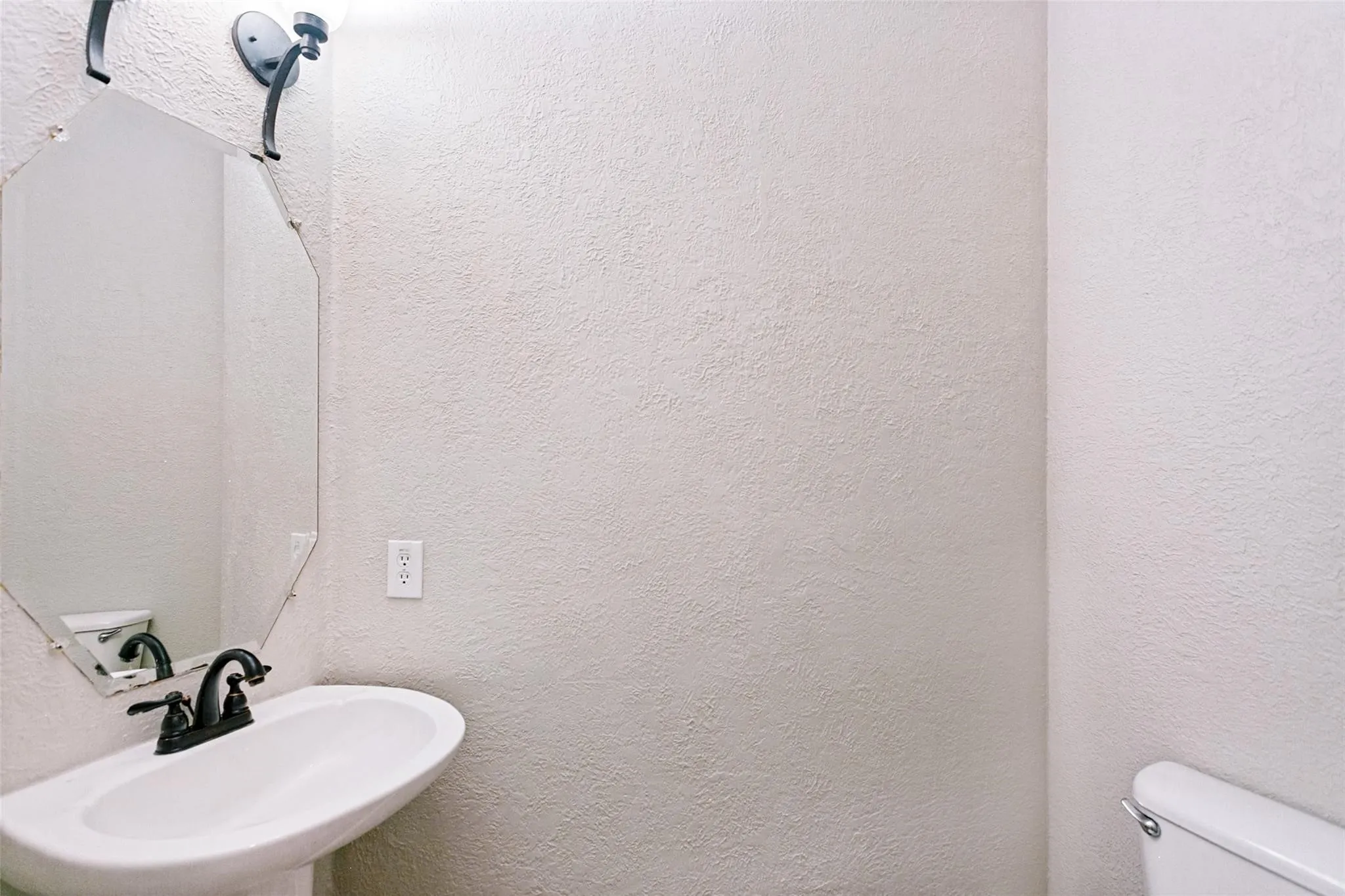 Pedestal sink with a dark bronze faucet, octagonal mirror, and wall-mounted light fixture
