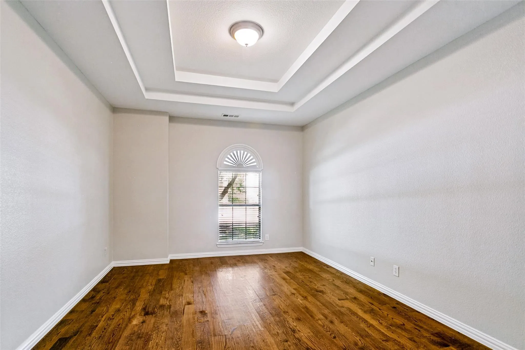 Tray ceiling with recessed lighting, wood-finish flooring, and arched window with blinds