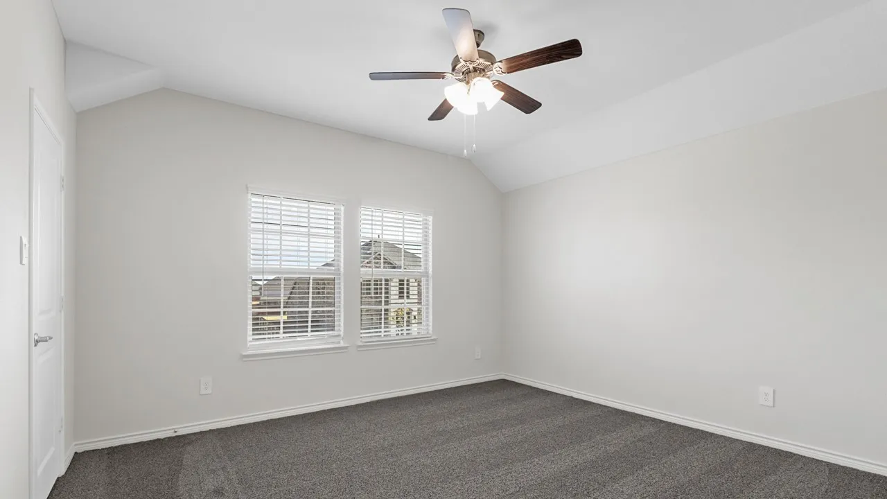 Vaulted ceiling, ceiling fan with integrated lighting, two windows with blinds, gray carpeting, and light-toned walls