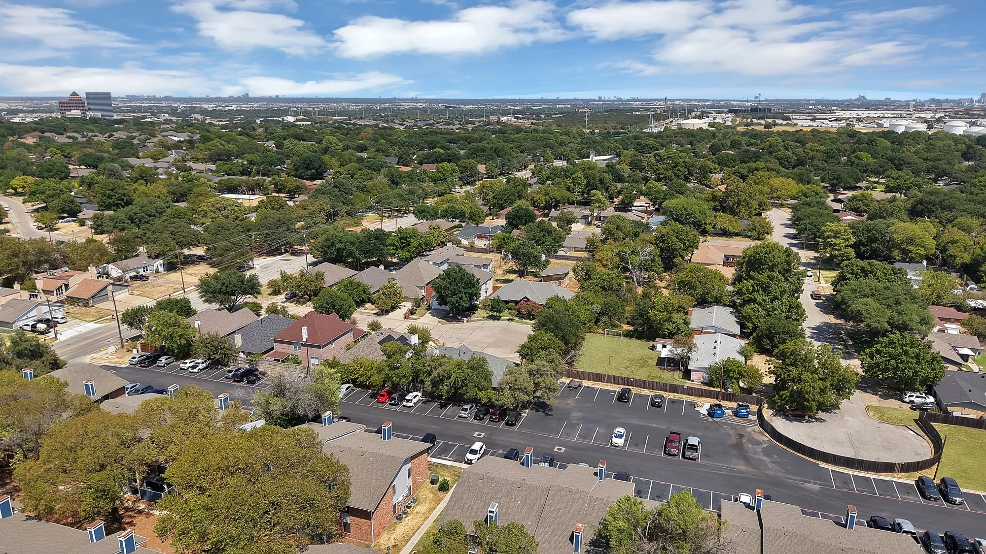 Extensive residential area featuring mature tree coverage, multiple paved parking areas, and distant city architecture