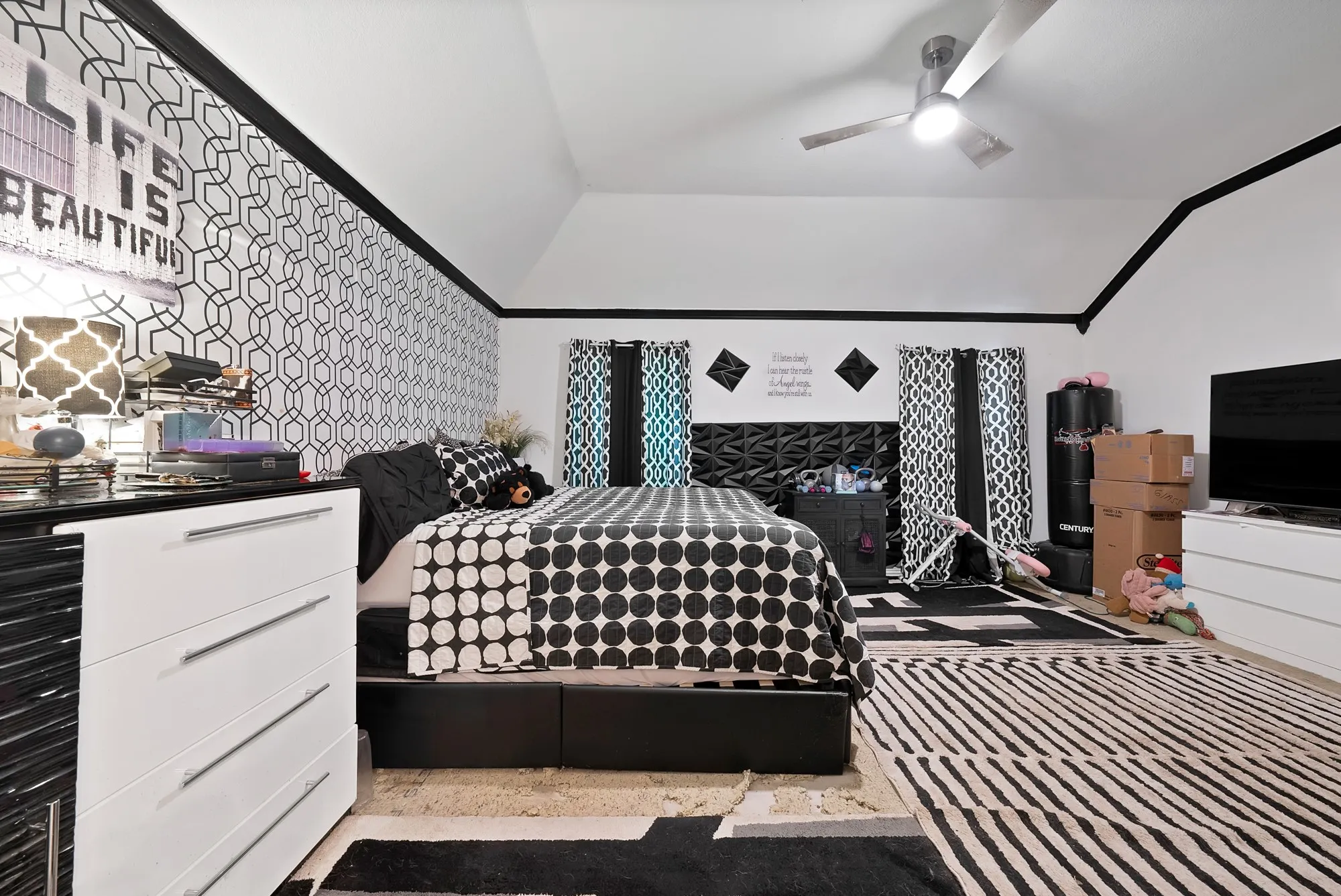Vaulted ceiling with a ceiling fan, accent wall with geometric pattern wallpaper, black trim detailing, and window with patterned curtains