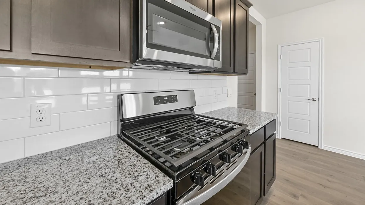 Stainless steel gas range with overhead microwave, white subway tile backsplash, dark wood-finish cabinetry, and speckled countertops