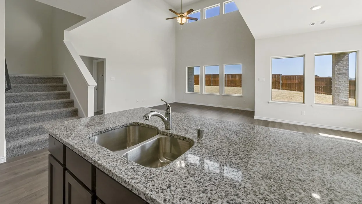 Kitchen island with a double basin stainless steel sink and polished stone countertop, open-concept design, vaulted ceiling, wood-finish flooring, and a staircase with carpeted treads