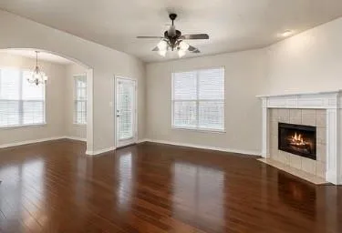 Dark wood-finish flooring, corner fireplace with tile surround, white mantel, ceiling fan with integrated lighting, and multiple windows with blinds