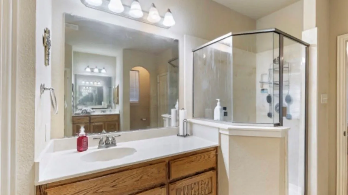 Wood-finish vanity with a light-toned countertop, a shower enclosure with clear glass panels, and a vanity-mounted light fixture with four sconces