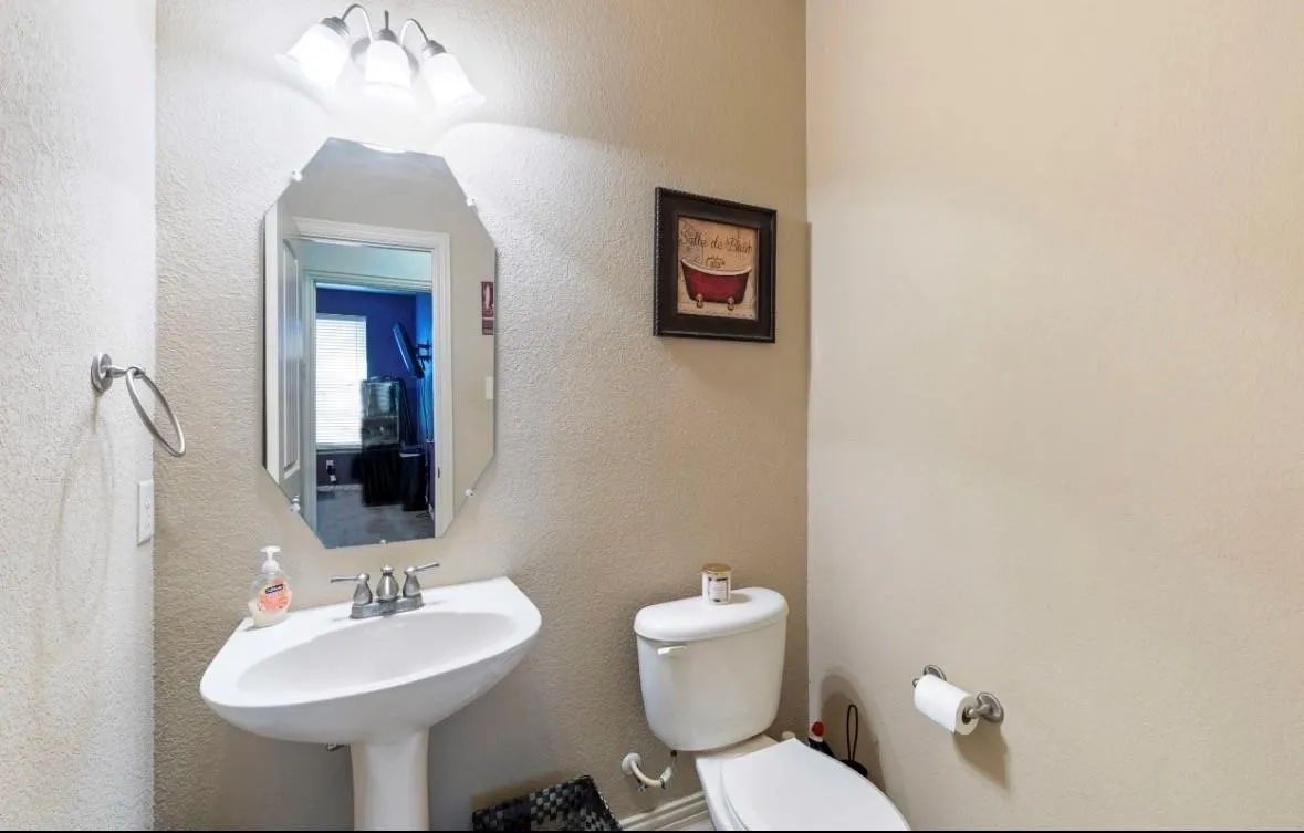 Compact half bath featuring a pedestal sink with chrome fixtures, wall-mounted mirror with integrated lighting, and a low-profile toilet
