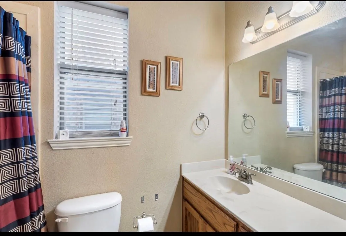 Bathroom featuring a single vanity with a white countertop and wood-finish cabinetry, a window with blinds, and a built-in mirror with overhead lighting