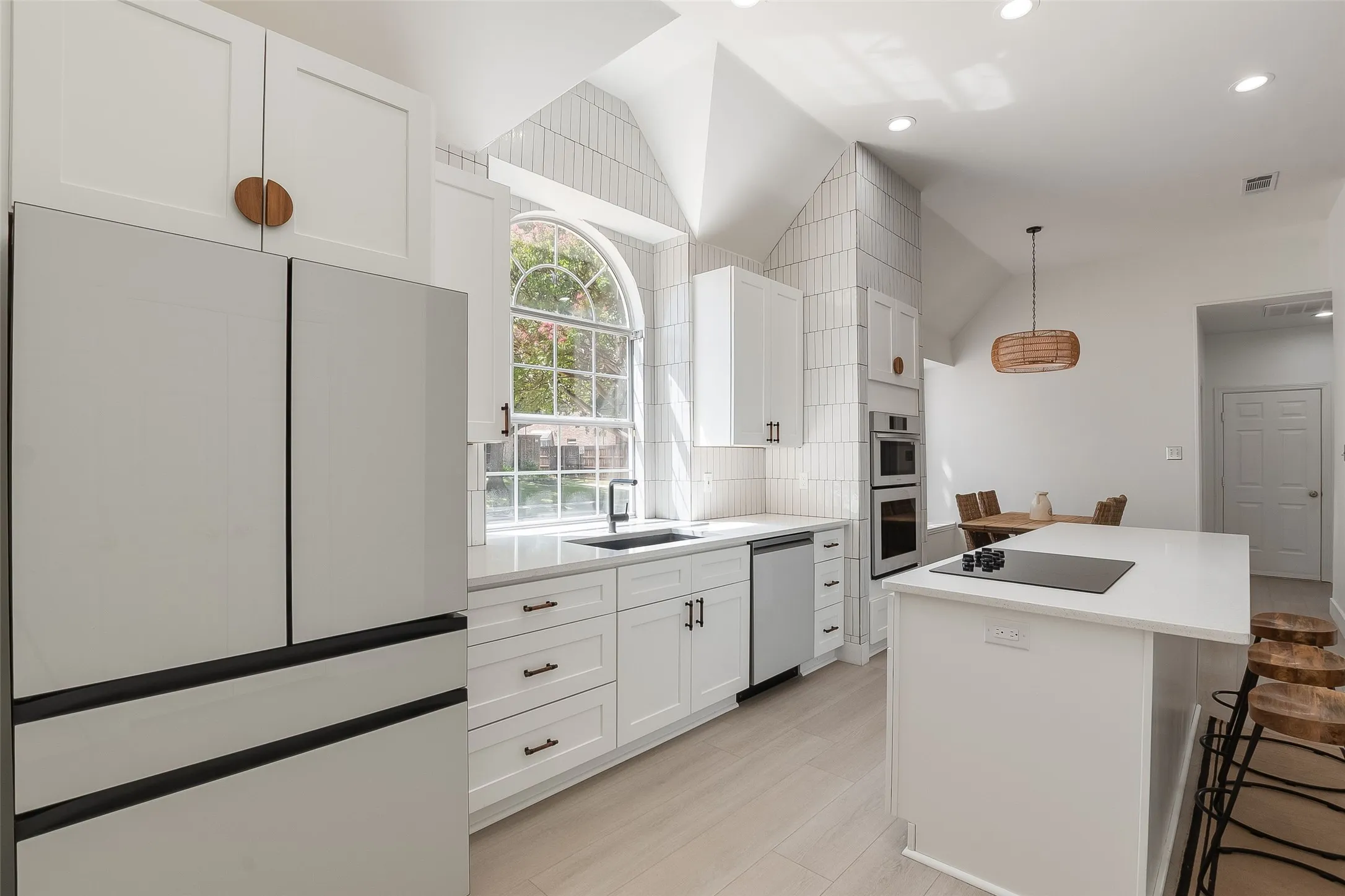 Vaulted ceiling, white cabinetry with dark hardware, built-in double oven, built-in cooktop on island, and arched window
