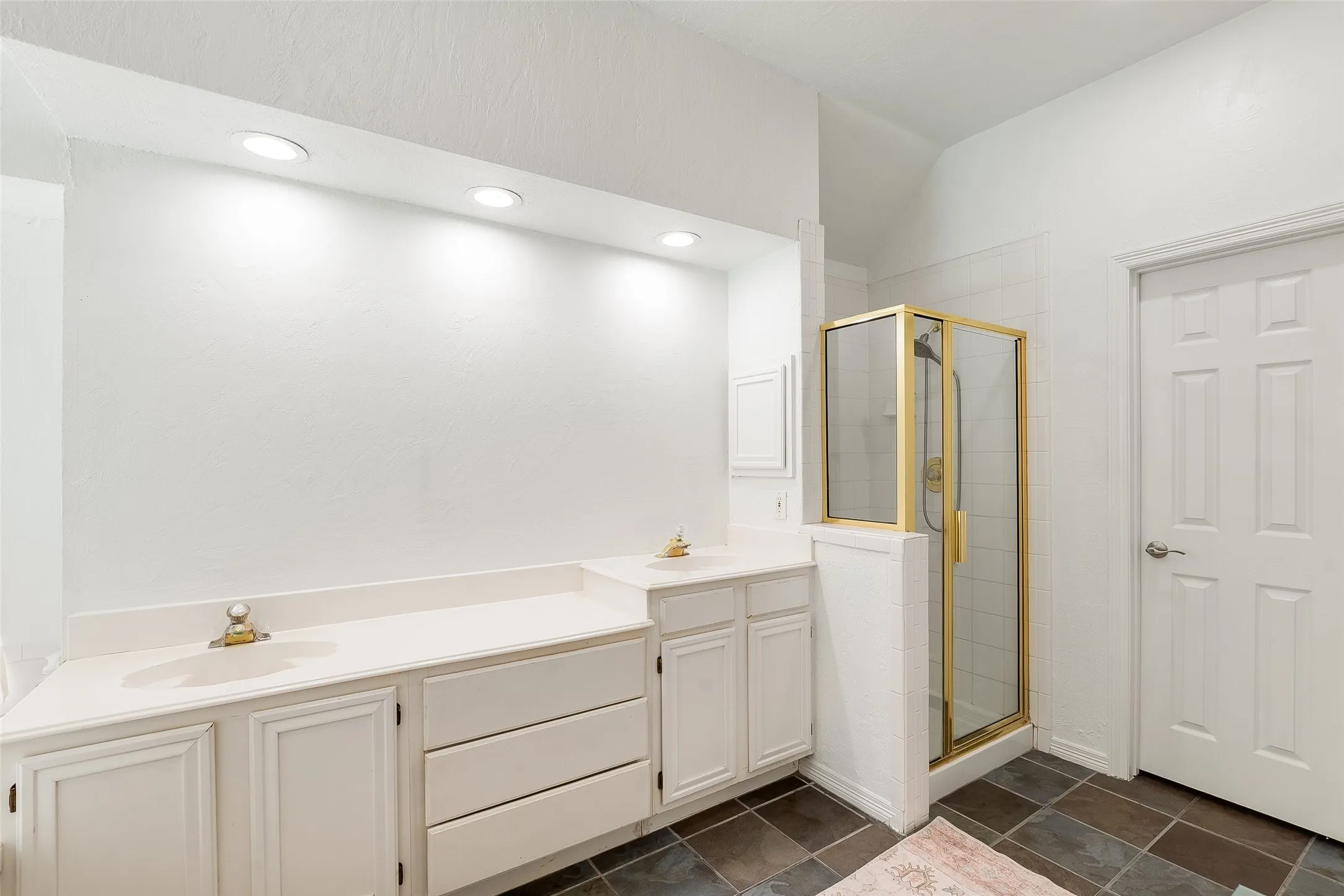 Recessed ceiling lighting, dual vanities with integrated sinks, white cabinetry, gold-framed shower enclosure, and dark square floor tiles