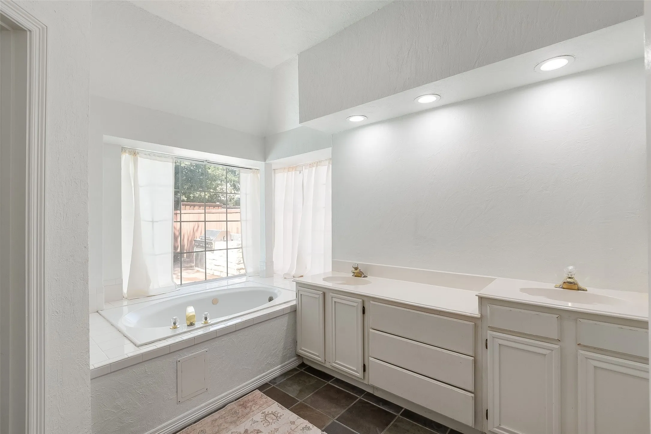 Spacious bathroom with an integrated soaking tub, dual vanity with solid surface countertops, recessed lighting, and dark tile flooring