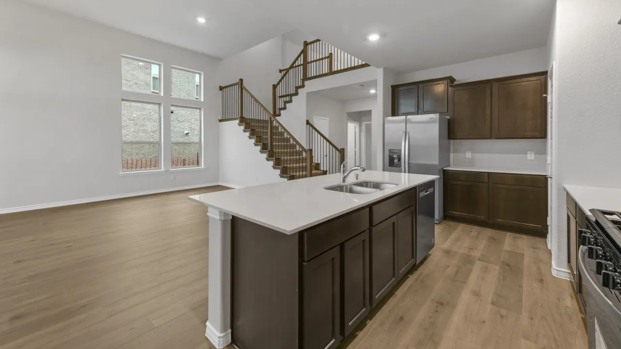 Open-concept living area with soaring ceilings, wood-finish flooring, a contemporary kitchen island featuring a double sink, stainless steel appliances, and a grand staircase with dark wood treads and wrought iron balusters