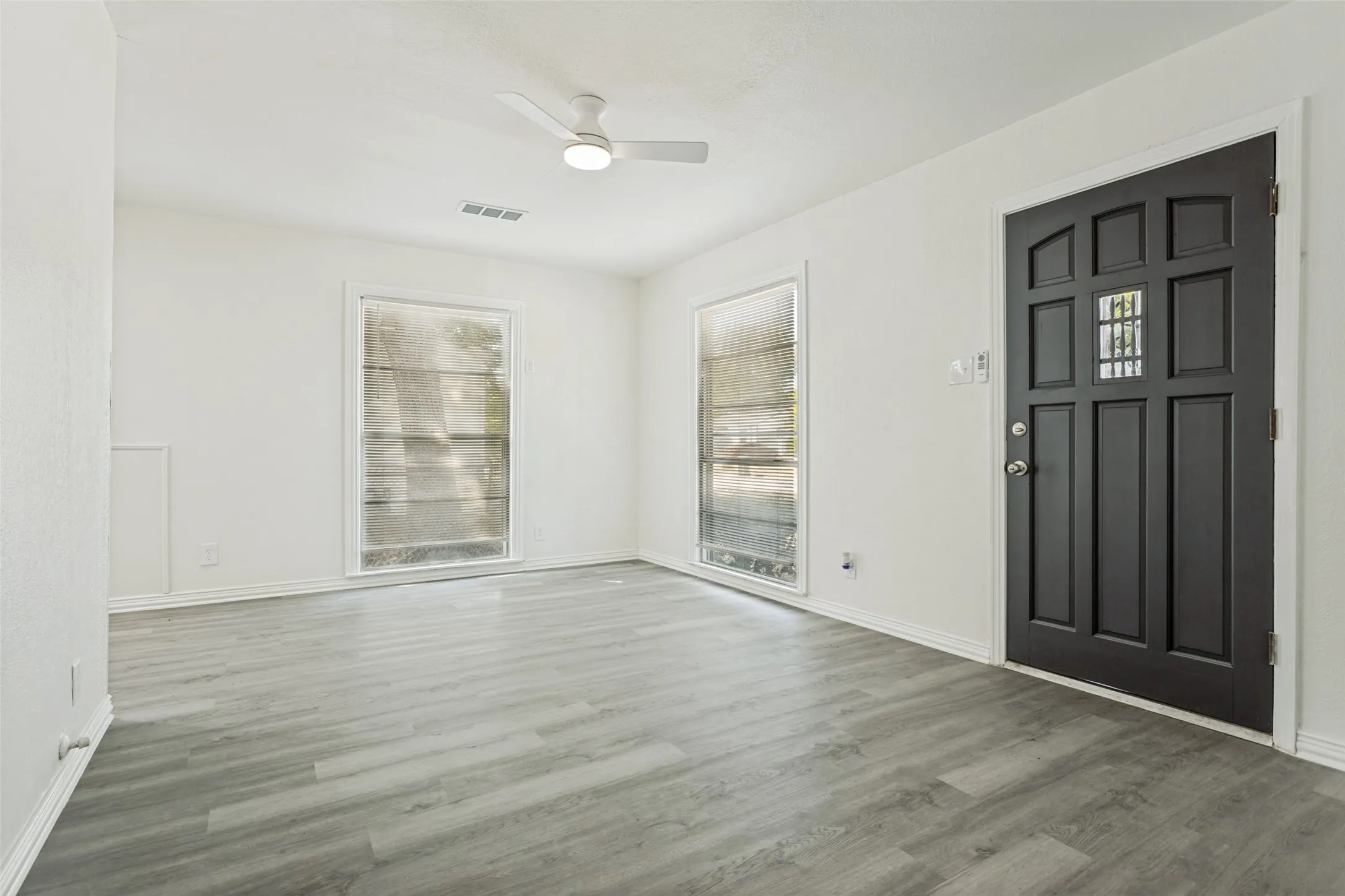 White interior walls, wood-finish flooring, two large windows, a ceiling fan, and a paneled entry door with decorative glass inserts