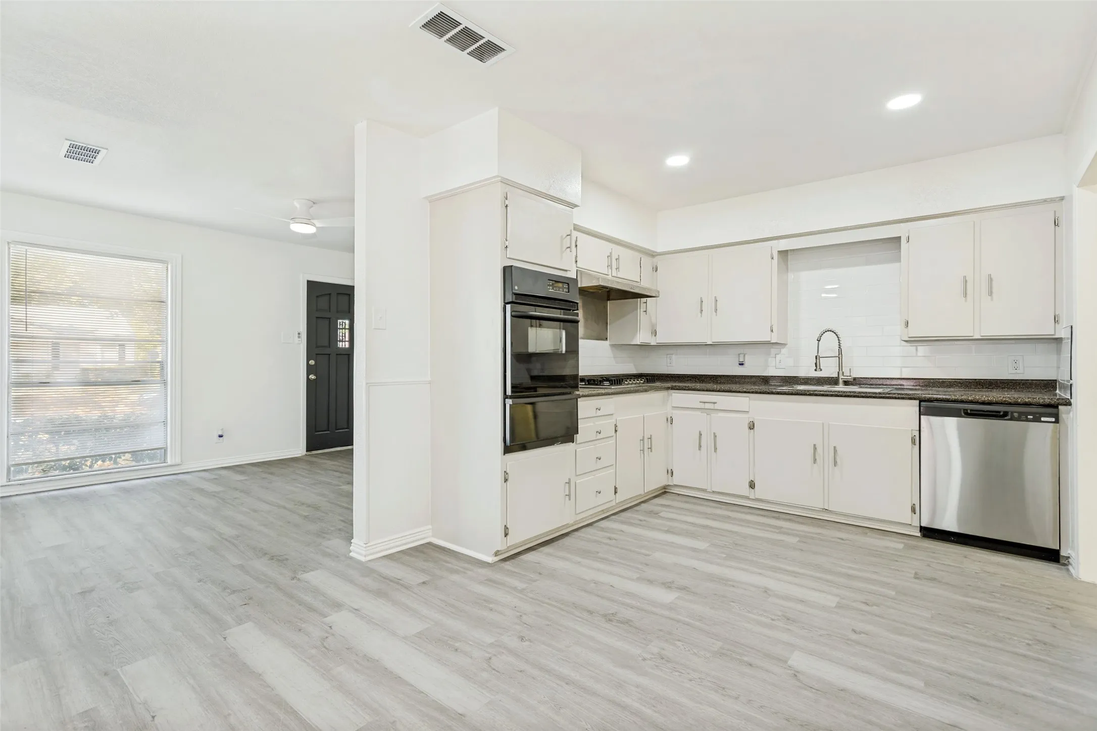 Light wood-finish flooring extending into the kitchen, white cabinetry, dark countertops, a stainless steel dishwasher, and a double wall oven