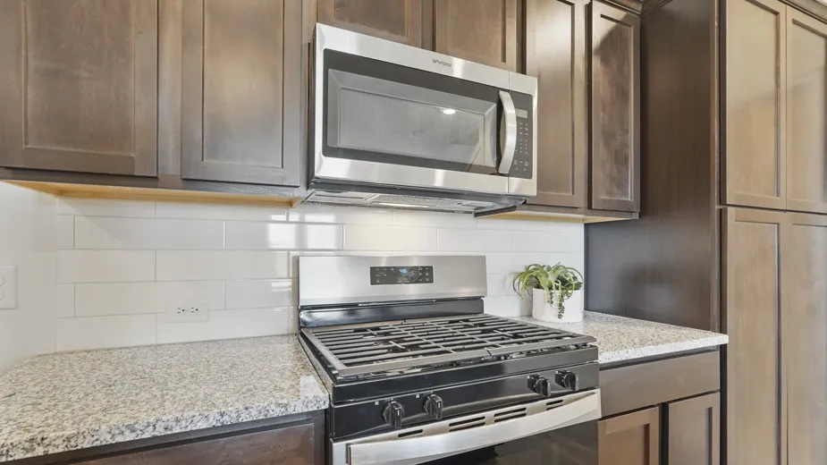 Stainless steel microwave, gas range, wood-finish cabinetry, granite-style countertops, and subway tile backsplash