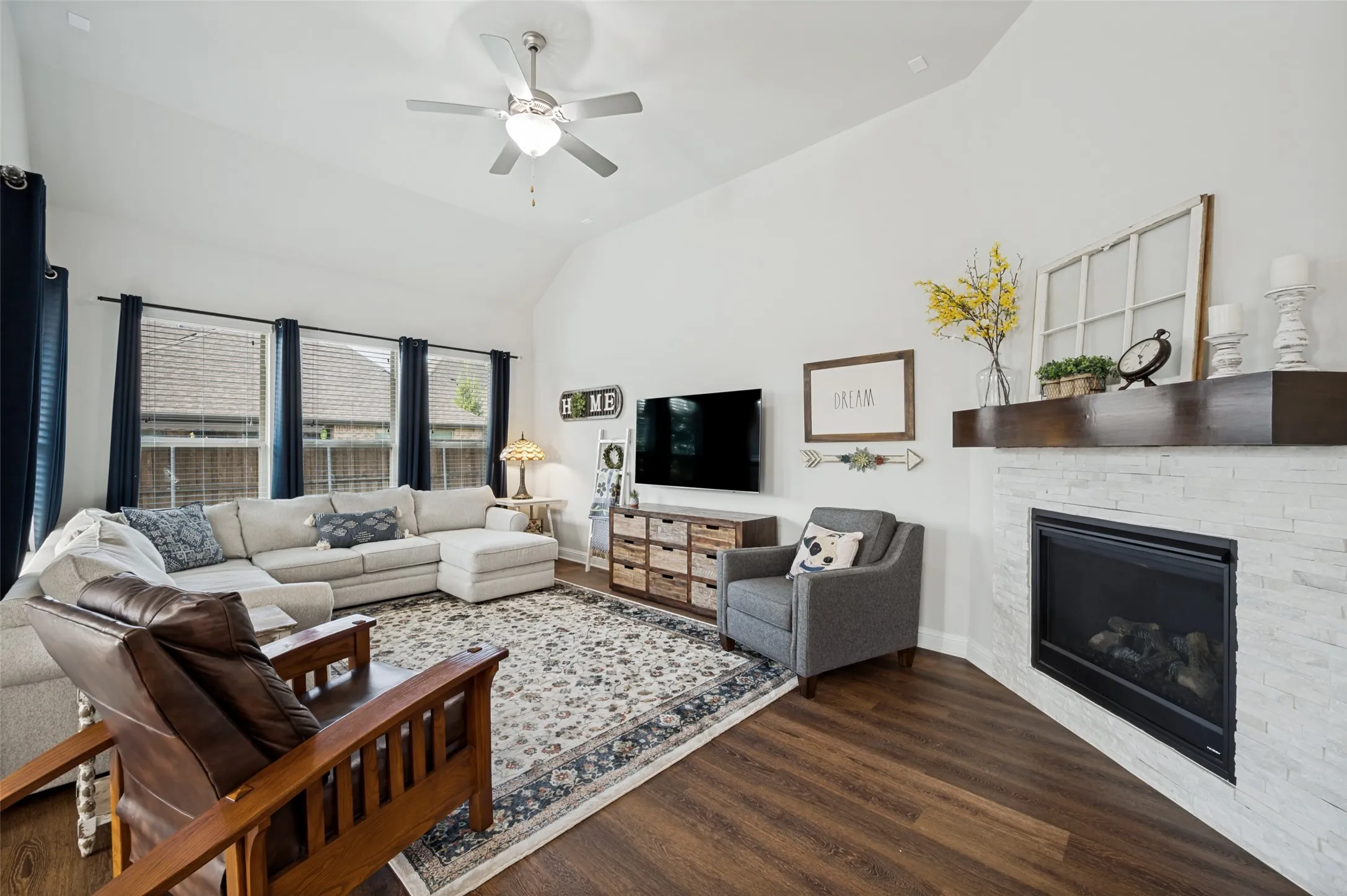 Spacious living room featuring vaulted ceilings, beautiful wood flooring, abundant natural light, and a striking stone fireplace with a wood mantel.