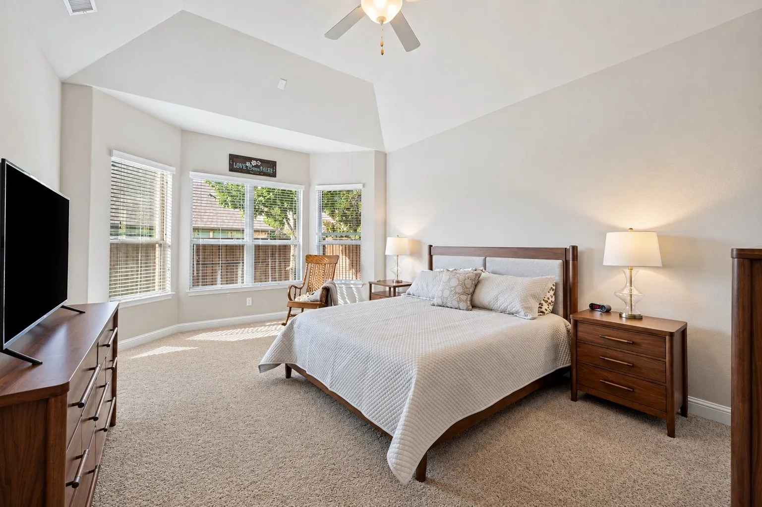 Spacious primary suite featuring a vaulted ceiling, plush carpeting, ceiling fan, and a wall of windows that fills the room with natural light.