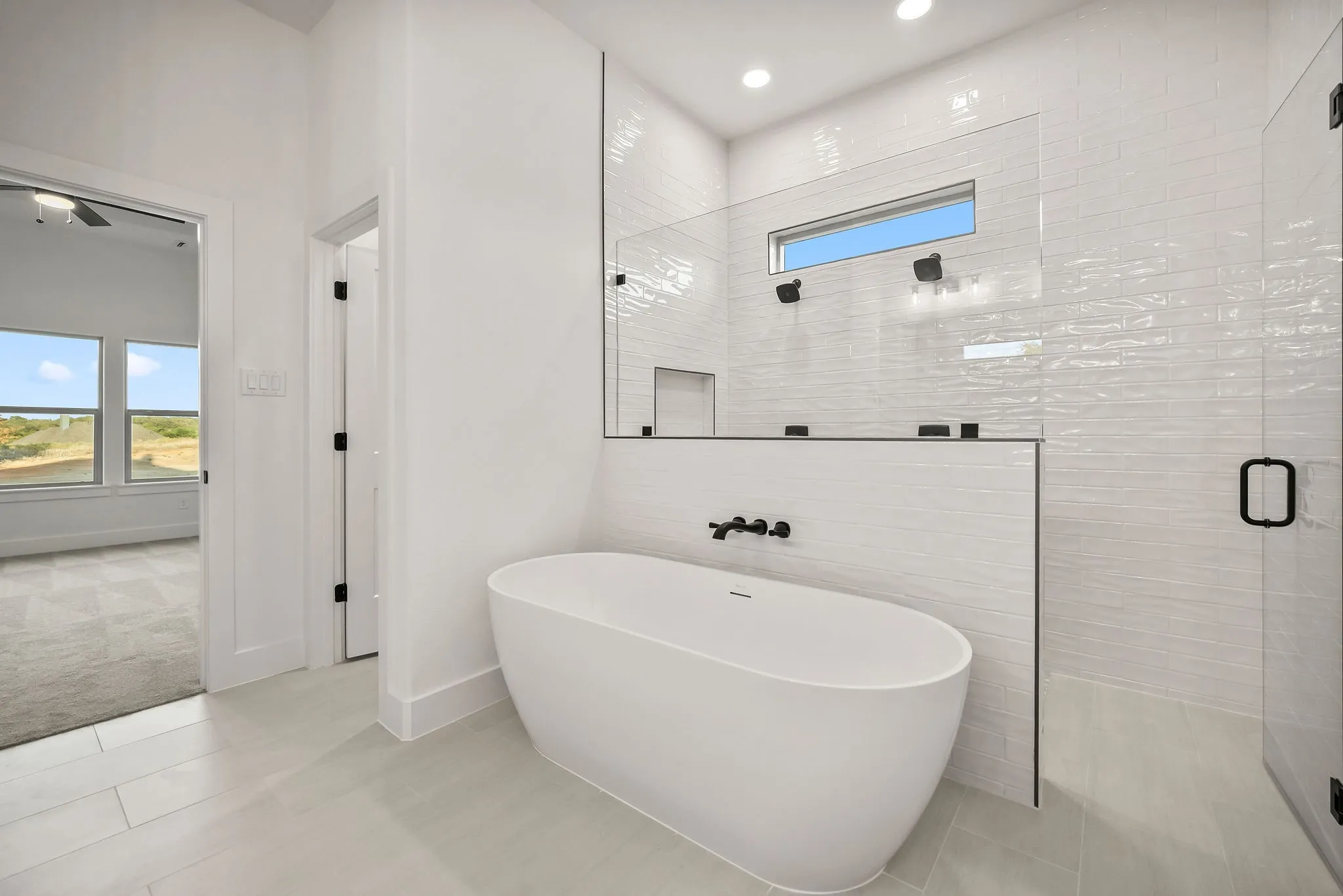 Freestanding soaking tub with matte black fixtures, walk-in shower with glass enclosure, tiled shower walls with horizontal accent window, and light-toned tile flooring