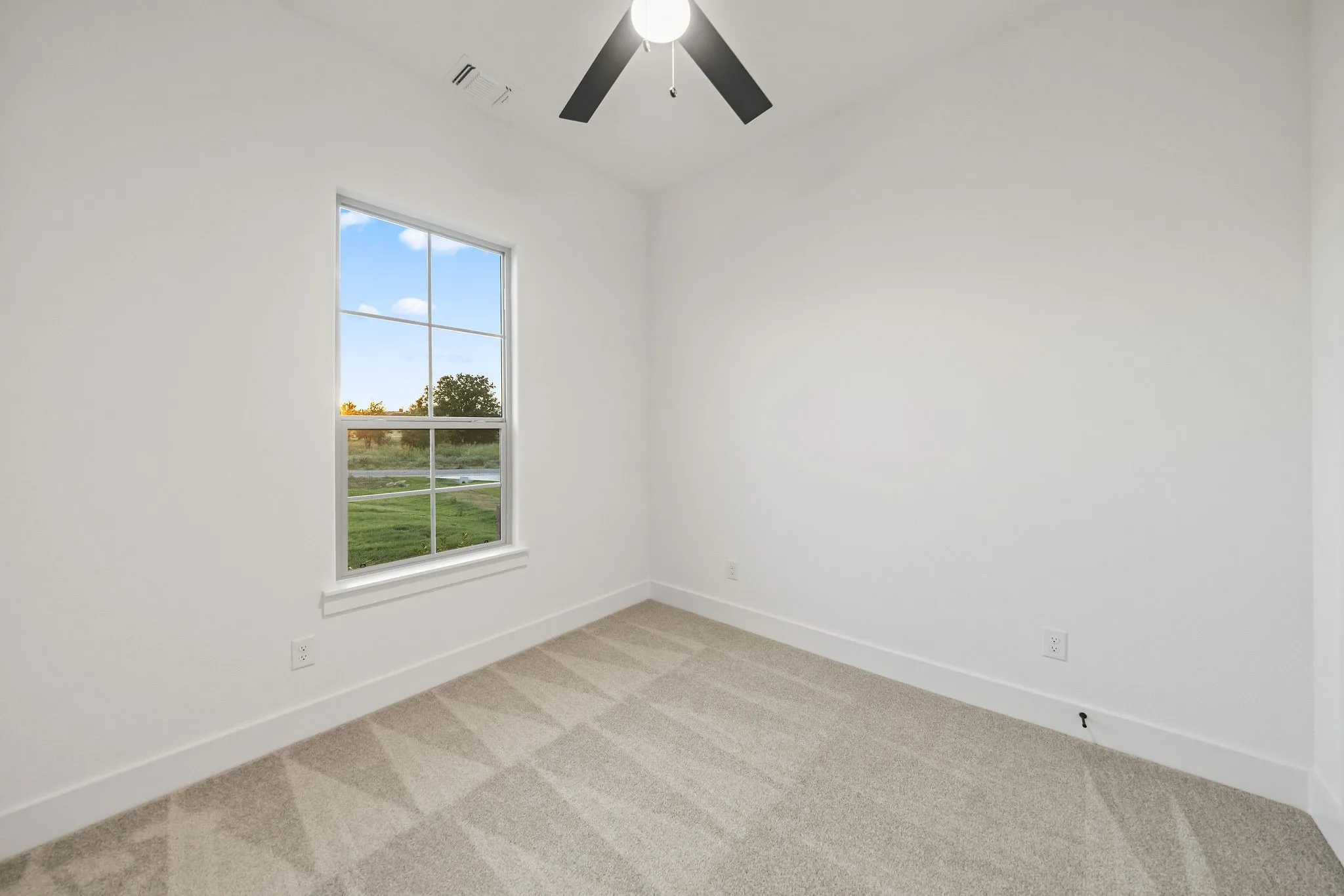 Window with outdoor scenery, ceiling fan with light fixture, light-toned walls, and patterned carpet flooring