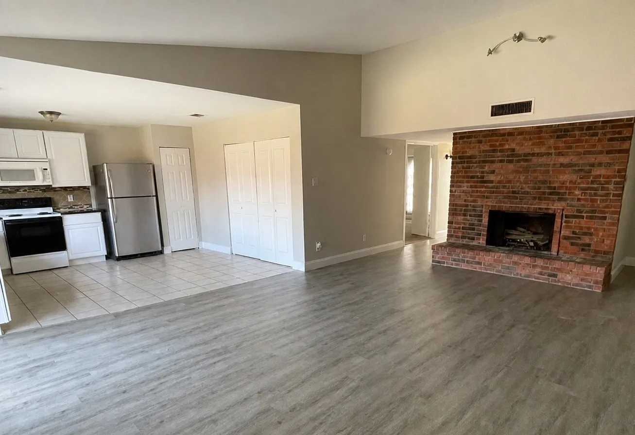 Vaulted ceiling enhancing the spacious living area, brick fireplace, wood-finish flooring, white cabinetry, and stainless steel appliances