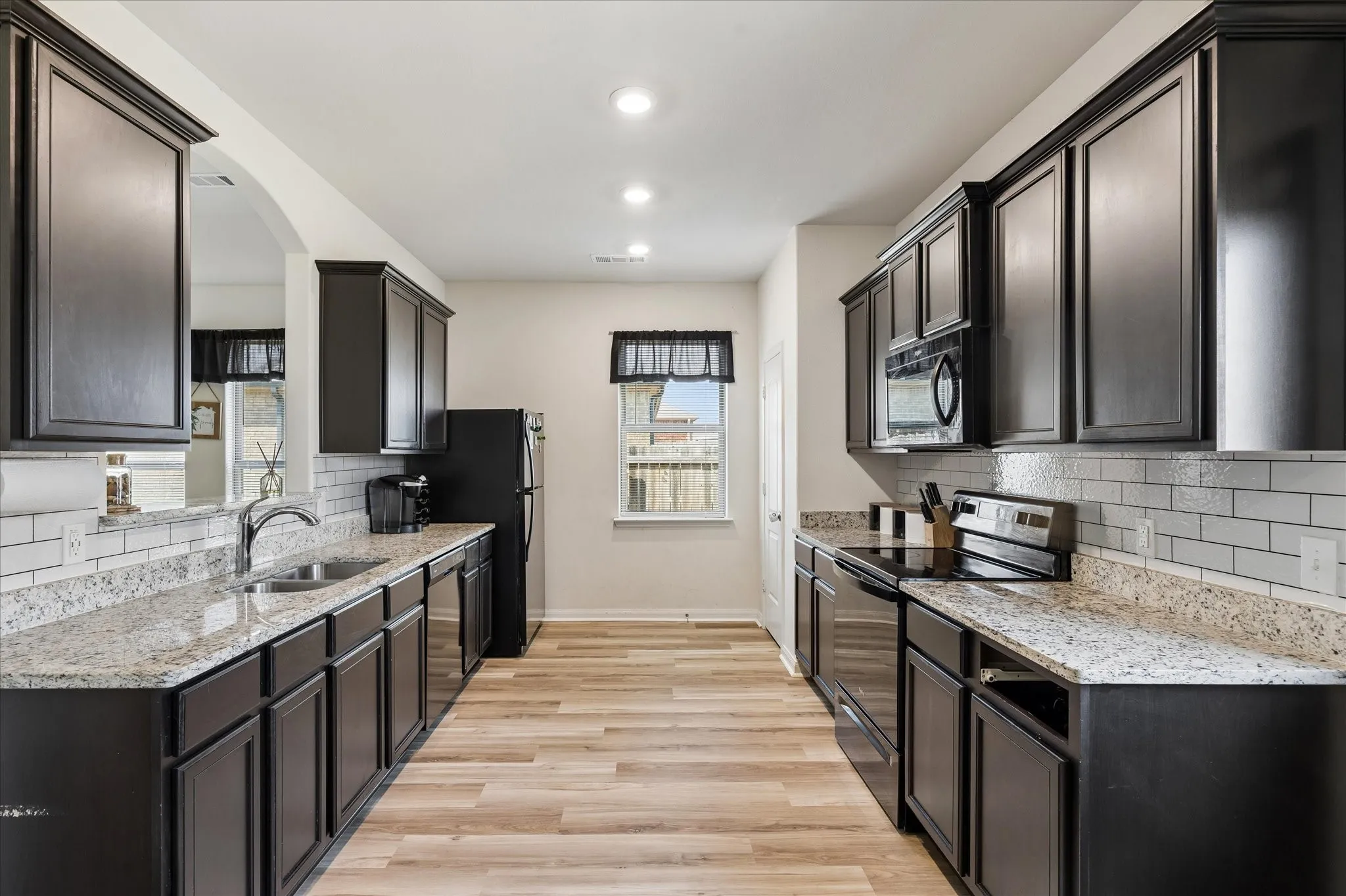 Galley kitchen featuring dark cabinetry, speckled countertops, stainless steel appliances, a subway tile backsplash, and light wood-finish flooring