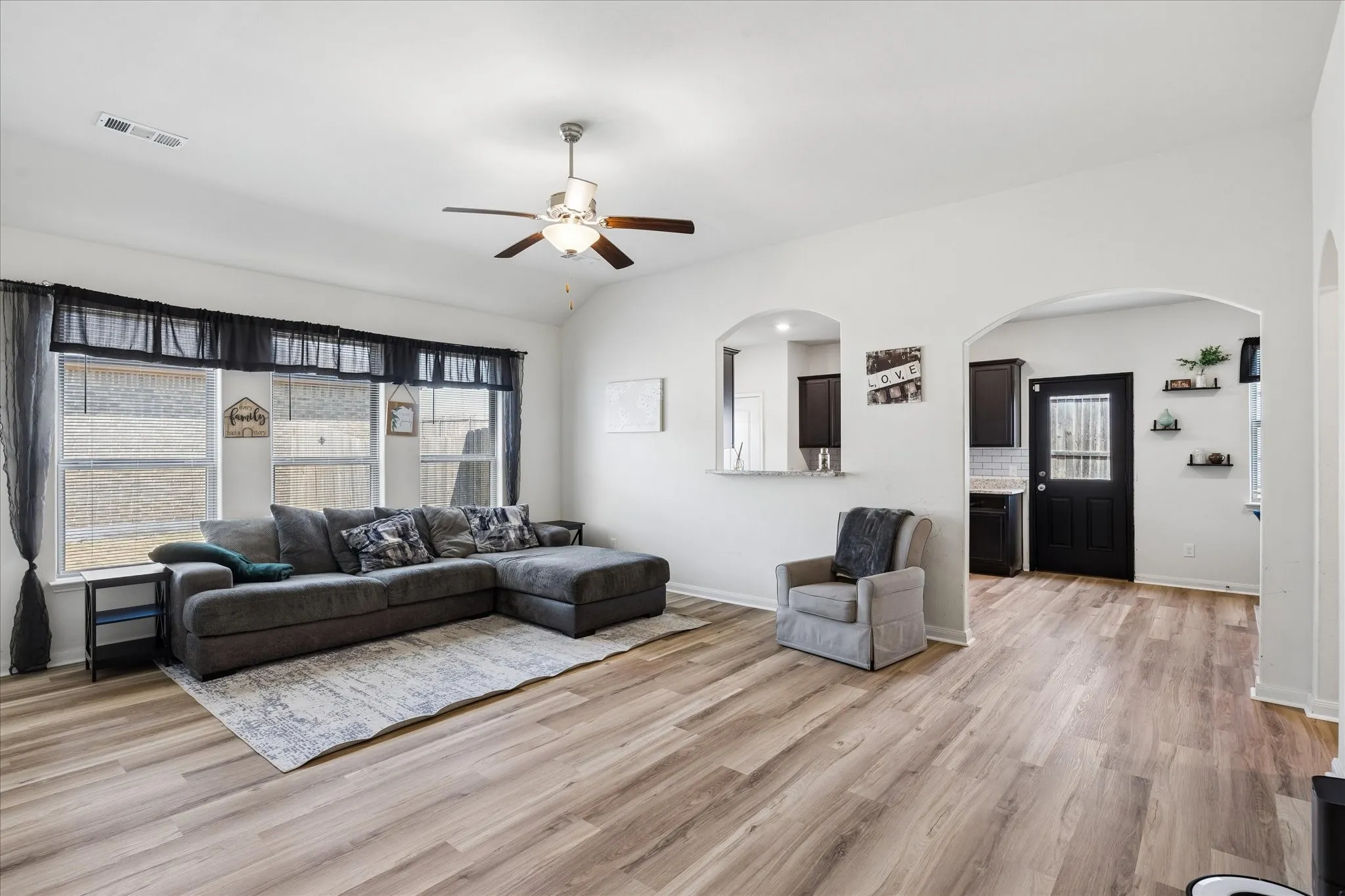 Spacious living area featuring light wood-finish flooring, a ceiling fan with integrated lighting, and multiple windows